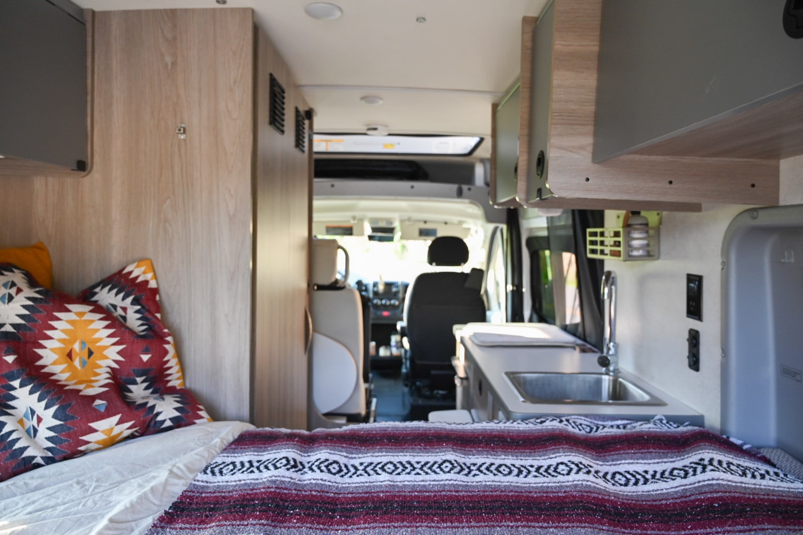 Inside a modern RV rental: a cozy bed with patterned blanket, compact kitchen sink, and view to the driver's cabin.. Winnebago Solis 59PX 2021