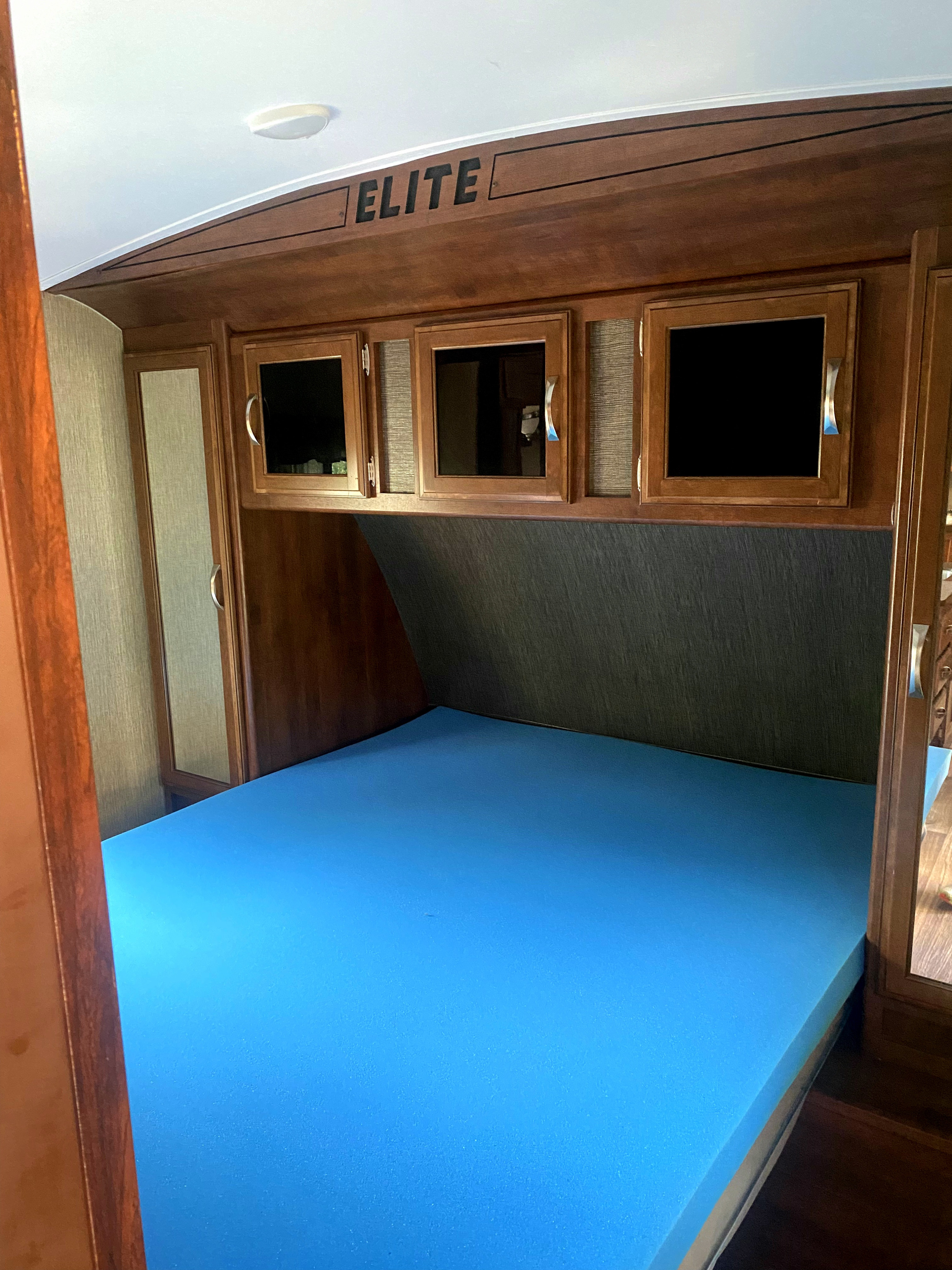 This RV rental bedroom features a blue foam mattress, dark wood cabinetry, and overhead storage labeled 'ELITE'.. Keystone Passport 2016