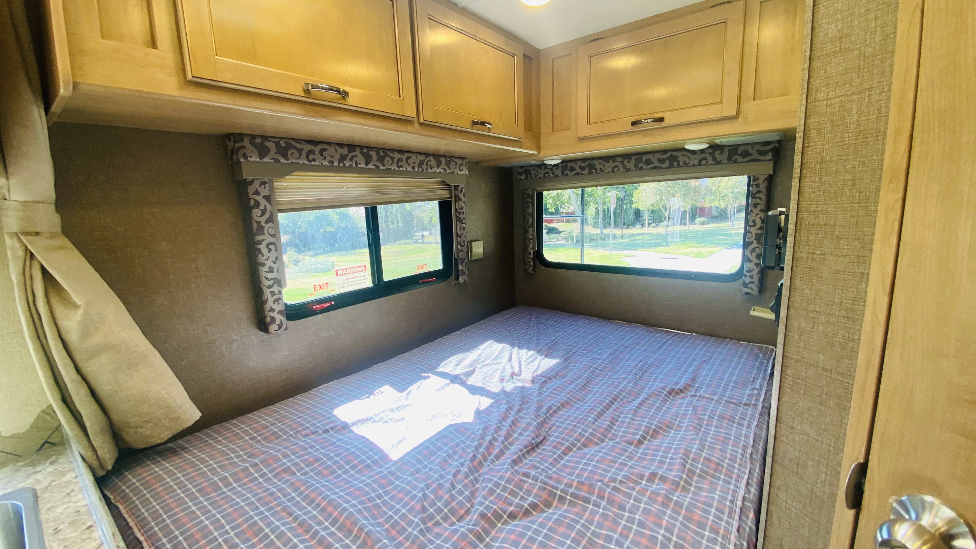 Inside an RV rental: a comfy bedroom area with a plaid bed, sunny windows, and wood cabinets.. Thor Four Winds 2019