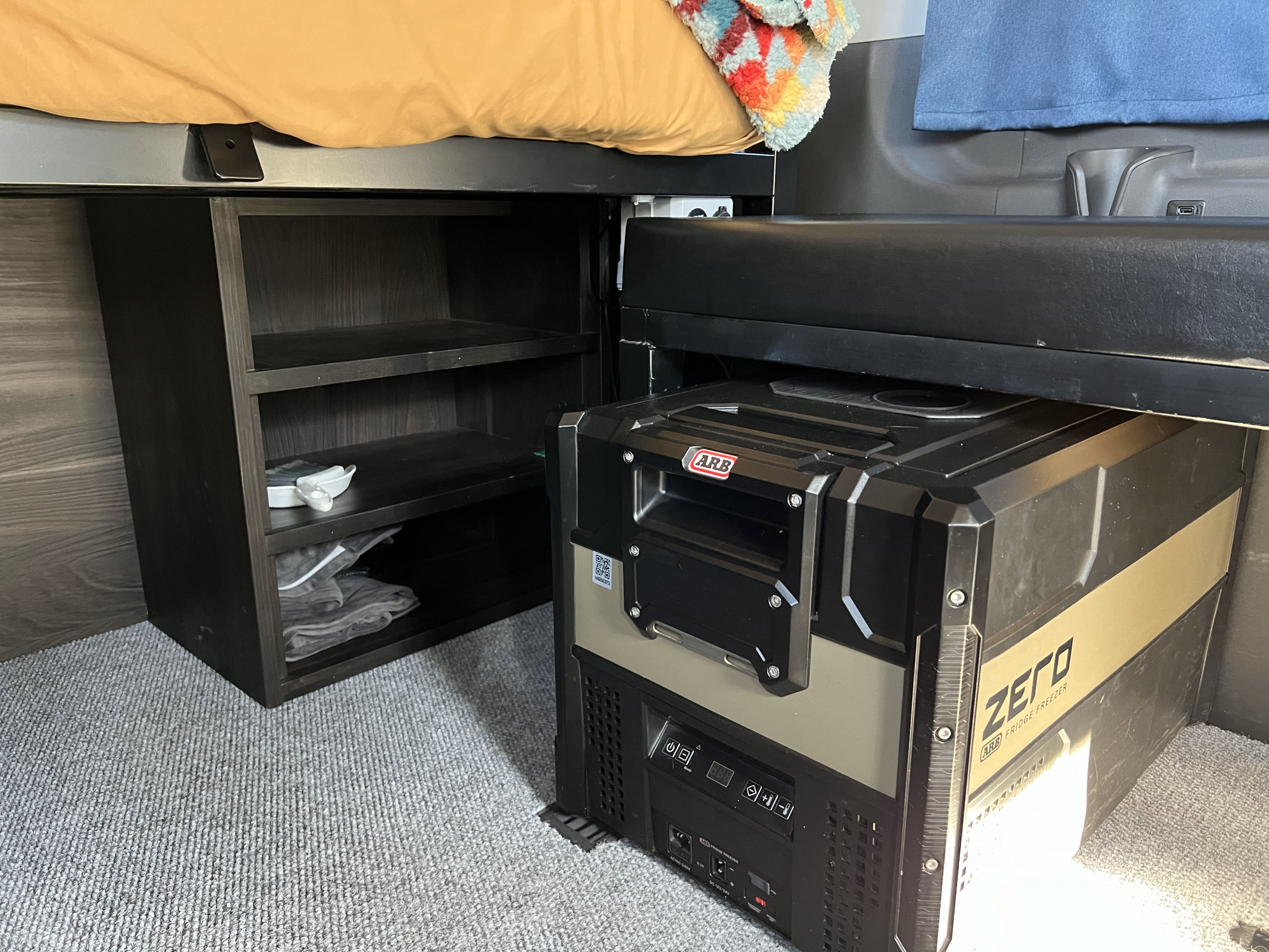 This rv rental interior features an ARB ZERO fridge freezer, dark storage shelves, and a bed, showcasing a compact living space.. Ford Transit 2020