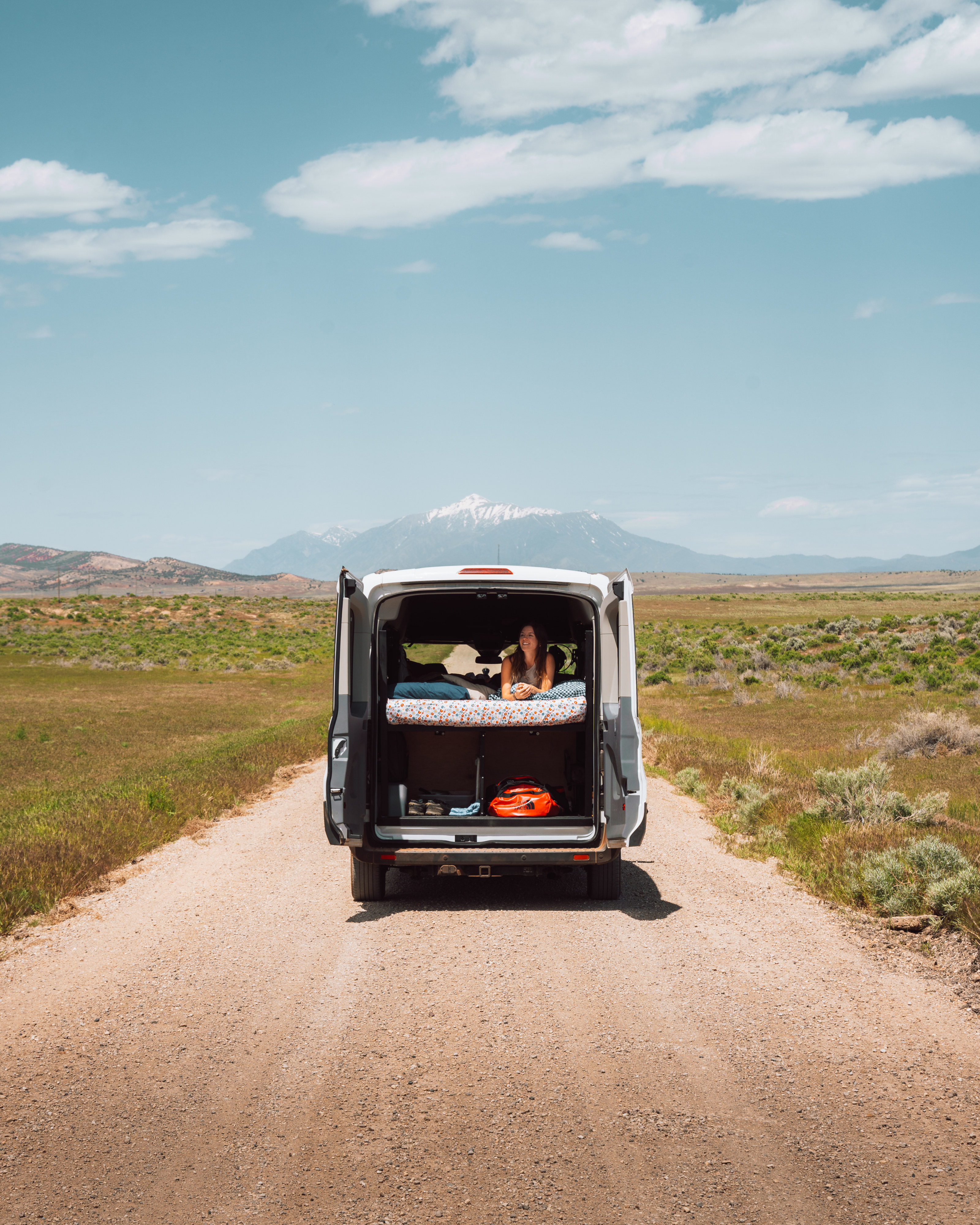 A woman relaxes in an RV rental van's open back, enjoying mountain views from her bed on a remote dirt road.. Ford transit 2019