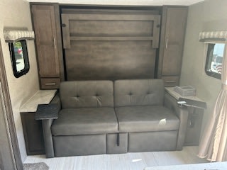 Inside this RV rental, a gray sofa sits below a folded Murphy bed, with wooden cabinets and counters on each side.. Forest River Flagstaff Micro Lite 2020