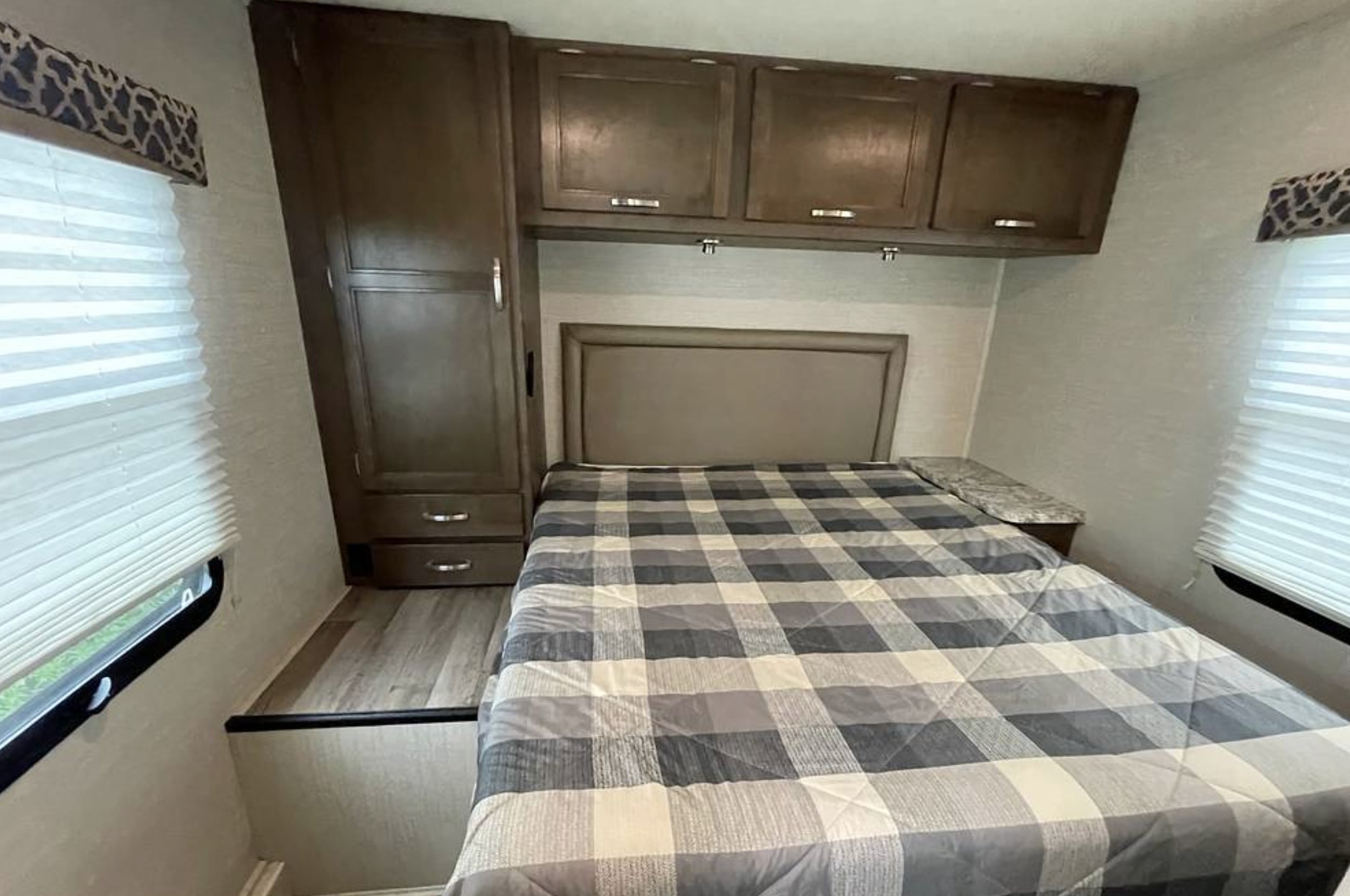 Cozy RV rental bedroom featuring a plaid bed, overhead cabinets, and bright windows. Ideal for comfortable travel.. Jayco Redhawk 2019