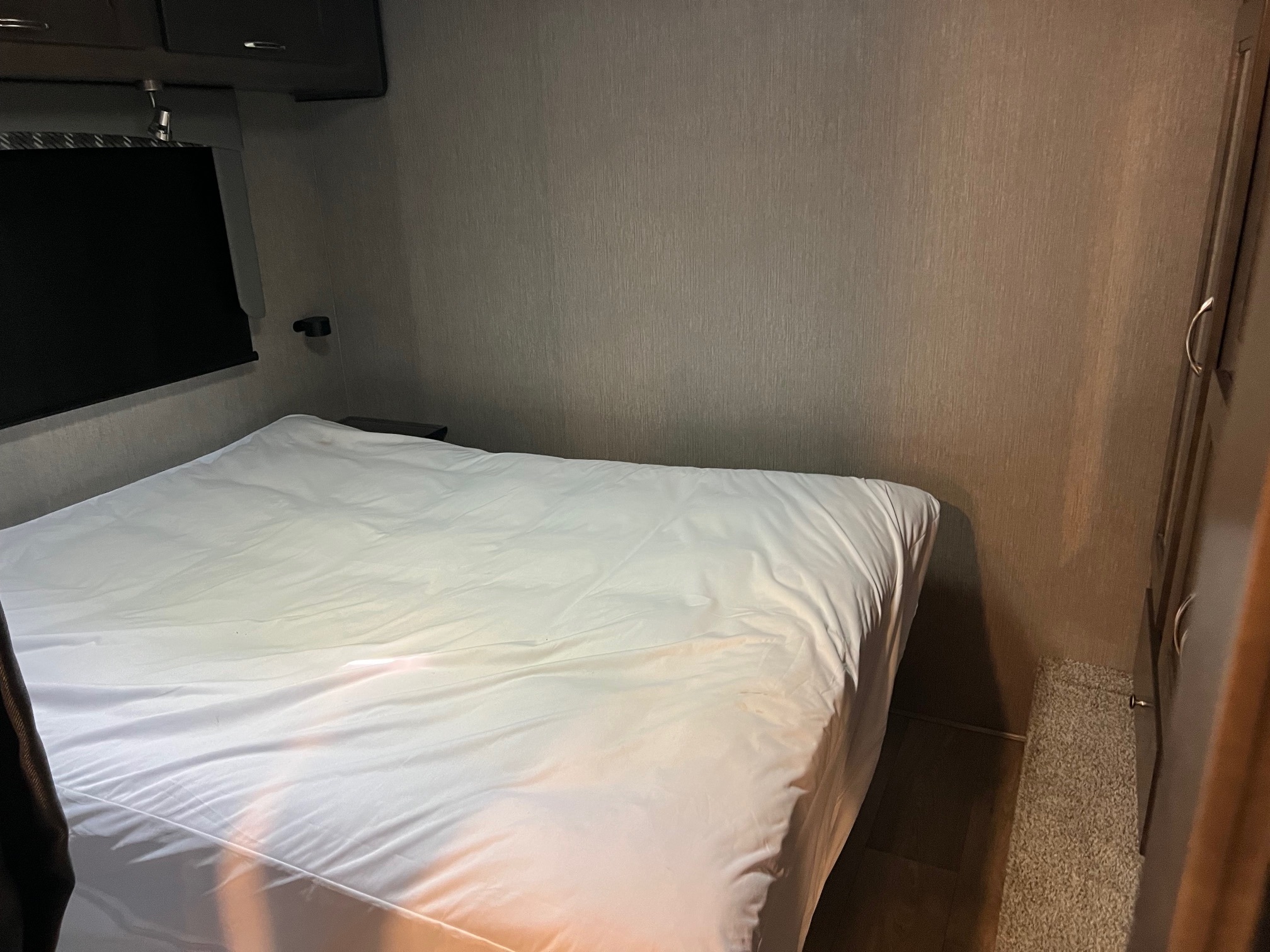 Cozy RV rental bedroom features a neatly made white bed, overhead storage, and a dark window blind. Ideal for travel.. Nexus RV Triumph 2023