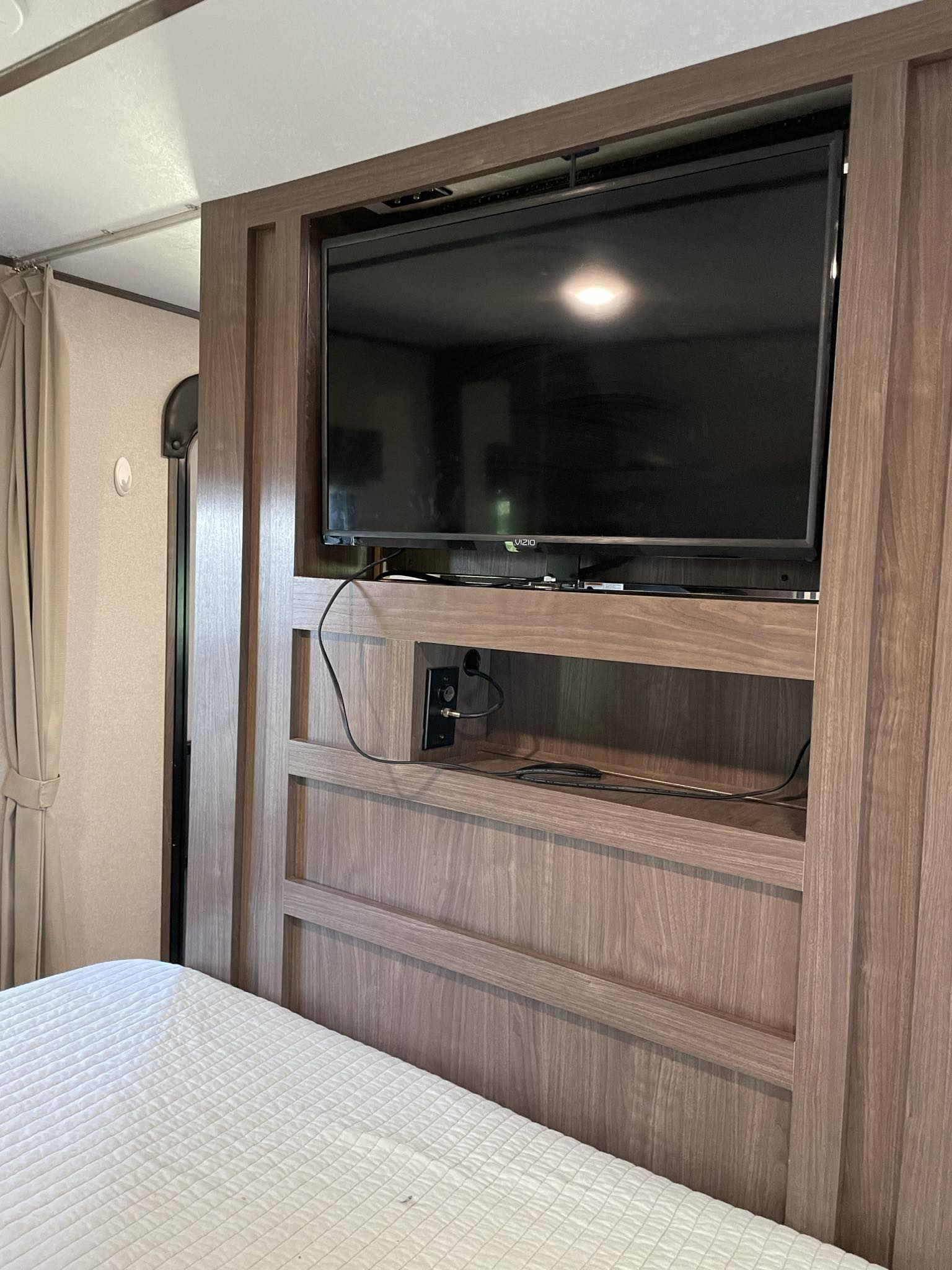 Cozy RV rental interior featuring a flat-screen TV mounted in wood-toned cabinetry above a storage shelf, beside a comfortable bed.. Keystone RV Hideout 2019