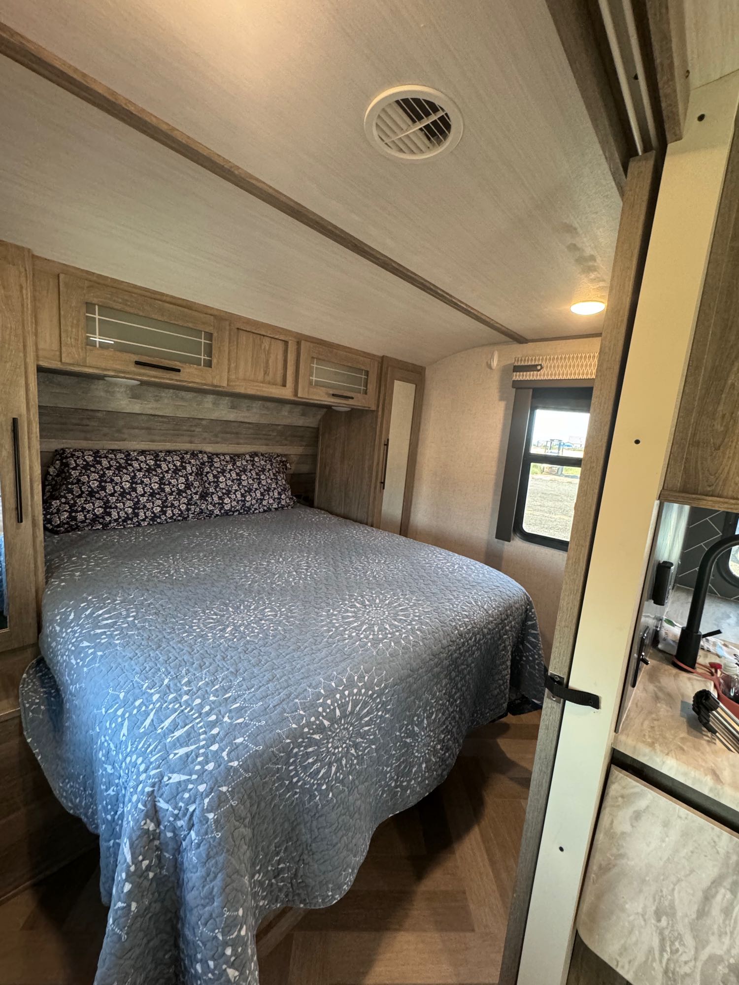 RV rental bedroom with a blue-quilted bed, overhead cabinets, and a window offers a comfortable travel stay.. Forest River Wildwood Heritage Glen HL 2021
