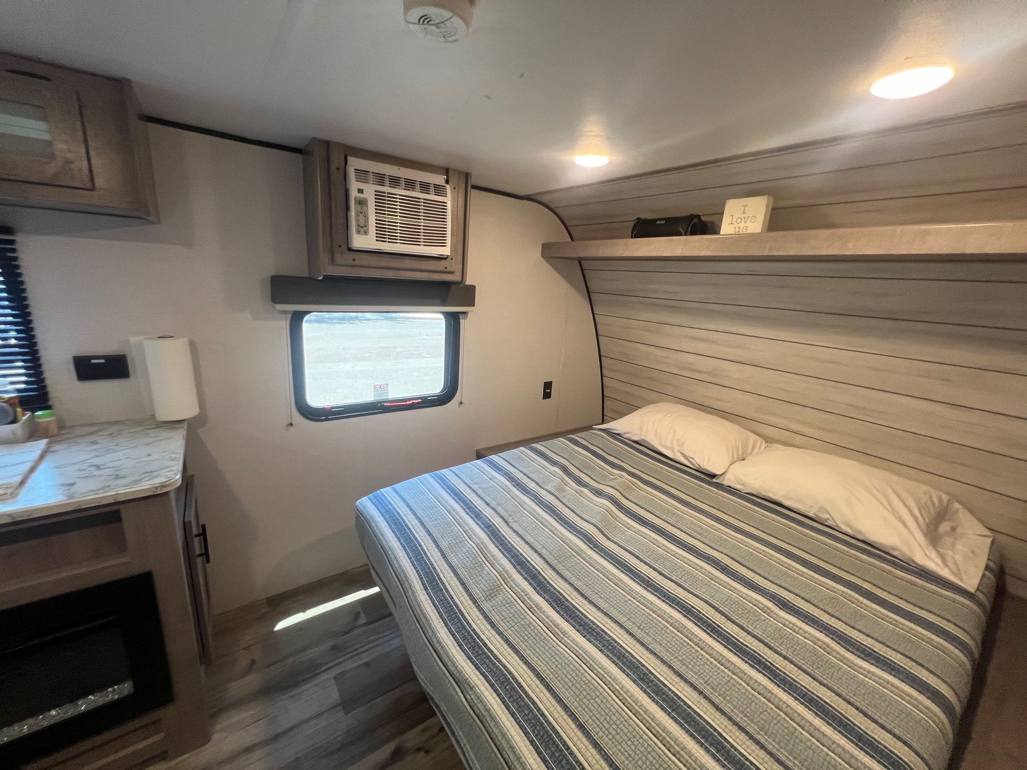 Inside an RV rental: a compact bedroom with a striped bed, window AC, overhead cabinets, and small kitchen counter.. Coleman Dutchman 2024