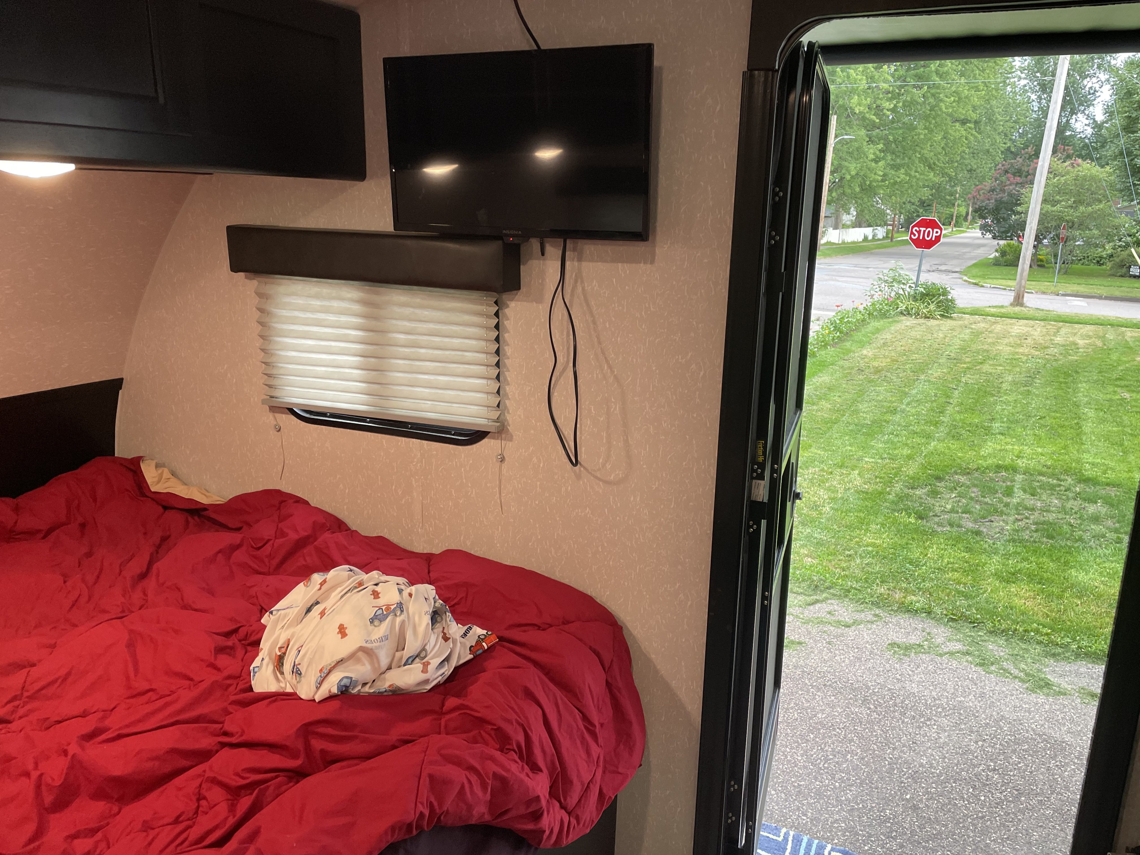 Cozy RV rental interior: red bed, mounted TV, open door revealing a green lawn, street, and stop sign outside.. Forest River Cherokee Wolf Pup 2022