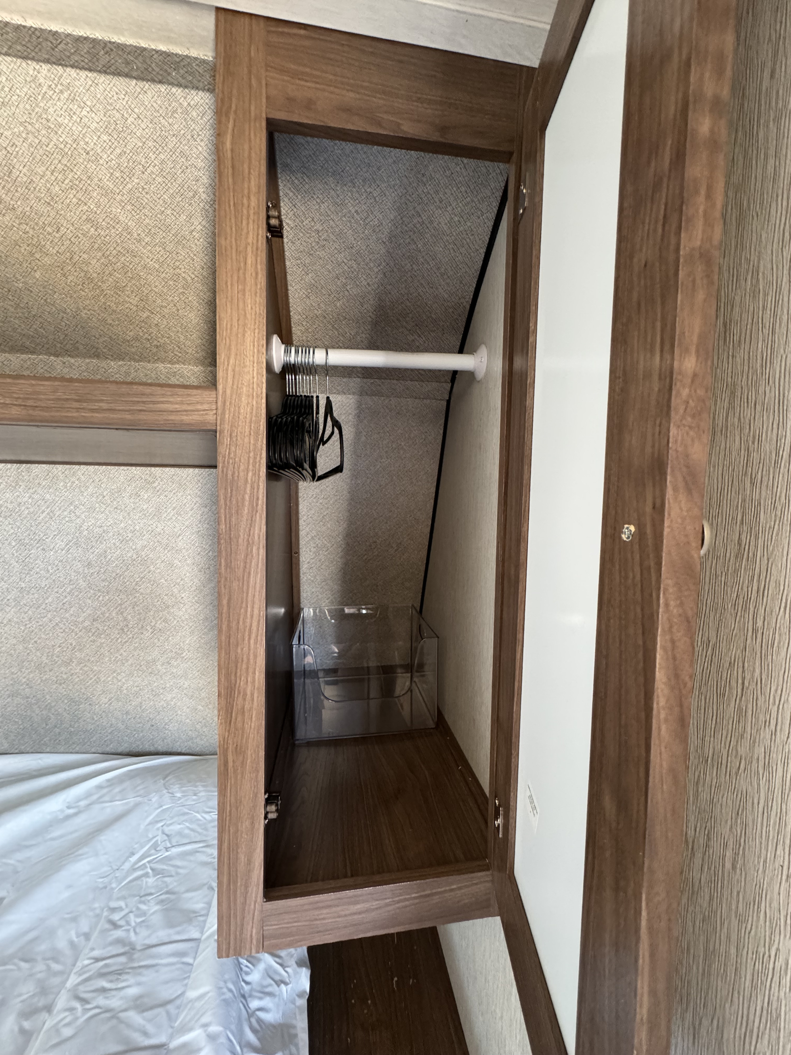 Open RV rental closet with a hanging rod, hangers, and a clear storage bin on the wood floor, next to a textured wall bed area.. Heartland RVs Trail Runner SLE 2019
