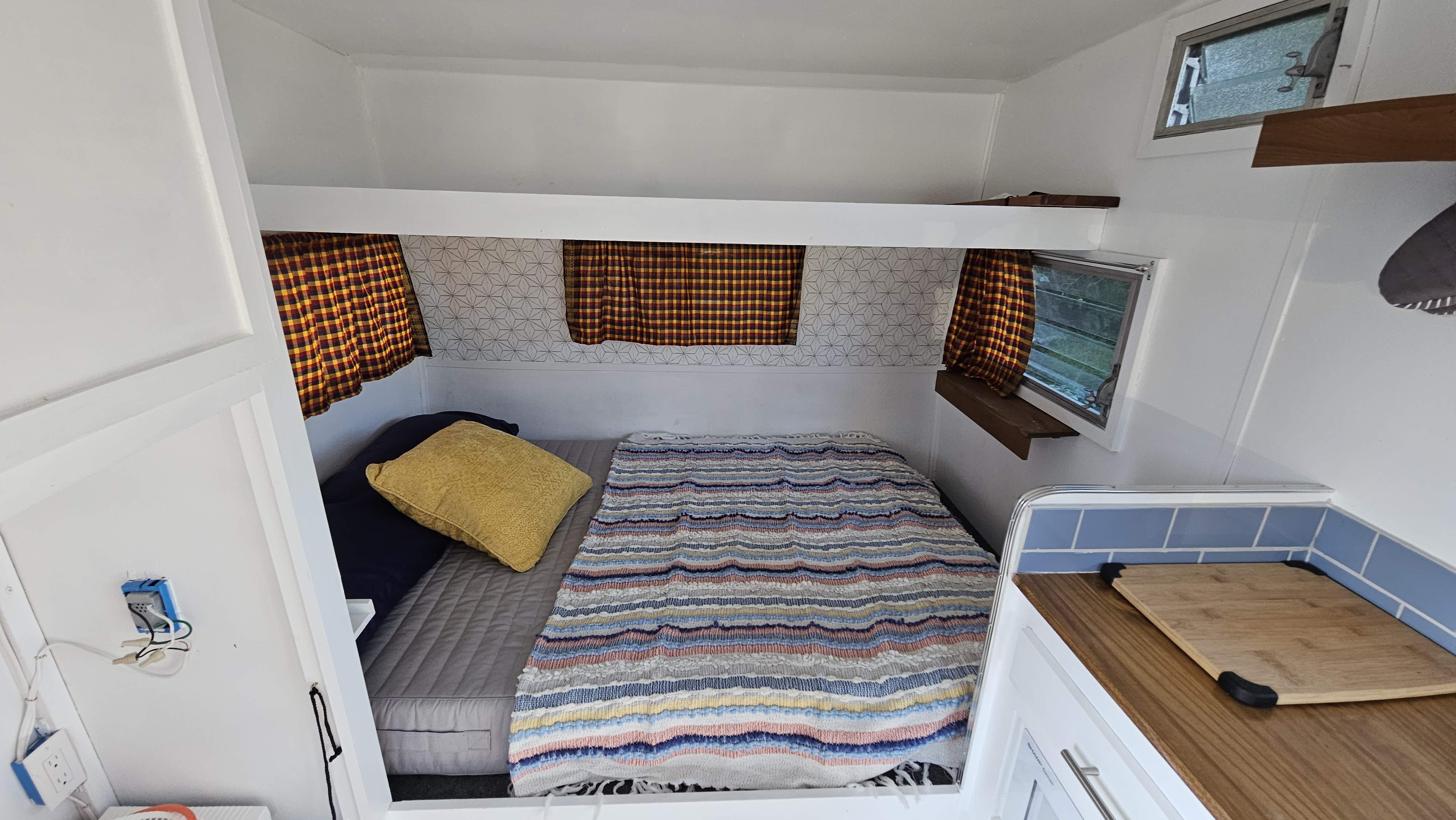 Inside a compact RV rental: a bed with striped blanket, plaid curtains, and a small kitchenette area.. Shasta Airflyte 1967