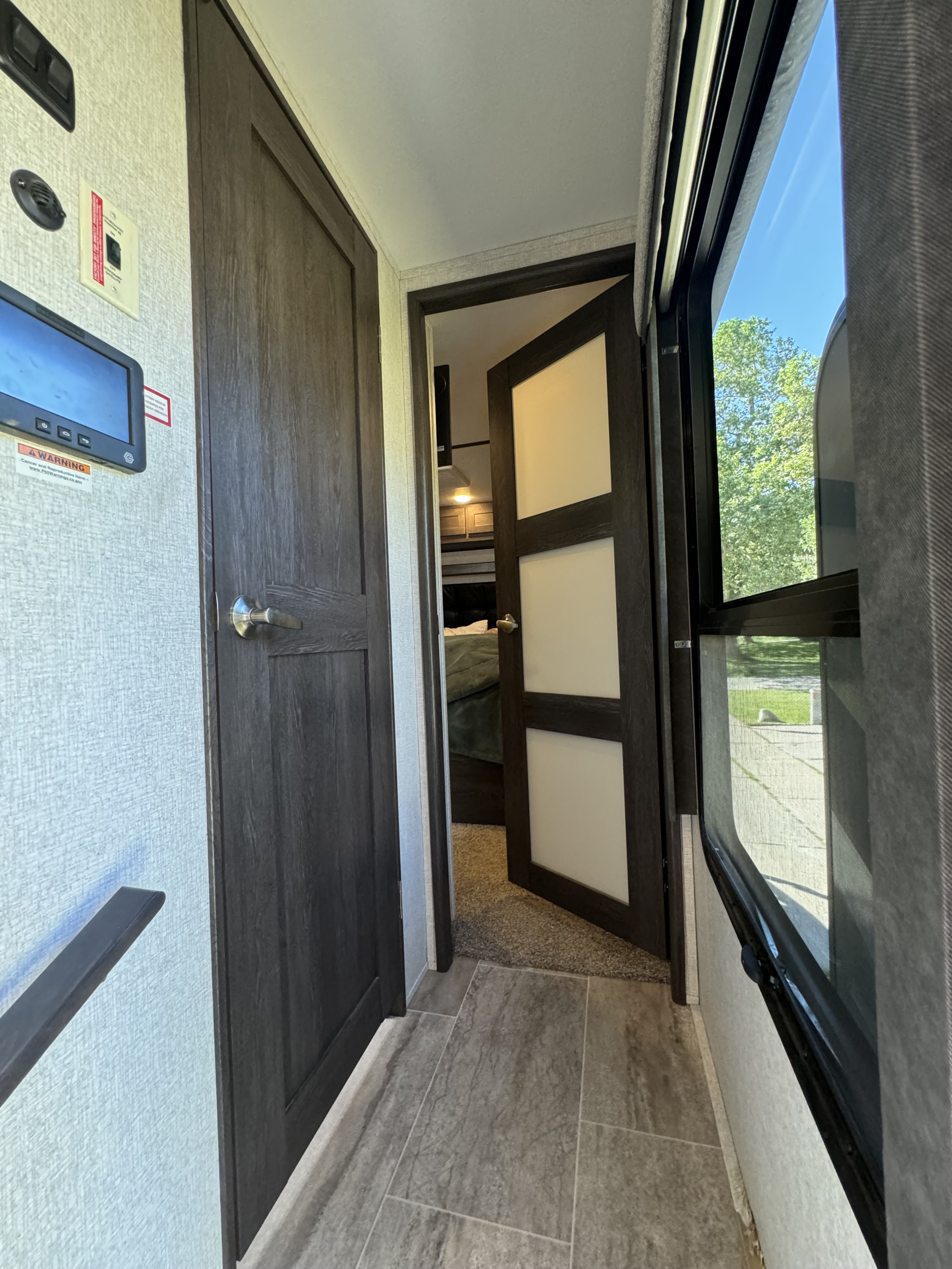 RV rental interior: compact passage with a control panel, dark wood doors, and window, leading to a private bedroom.. Keystone Raptor 2020