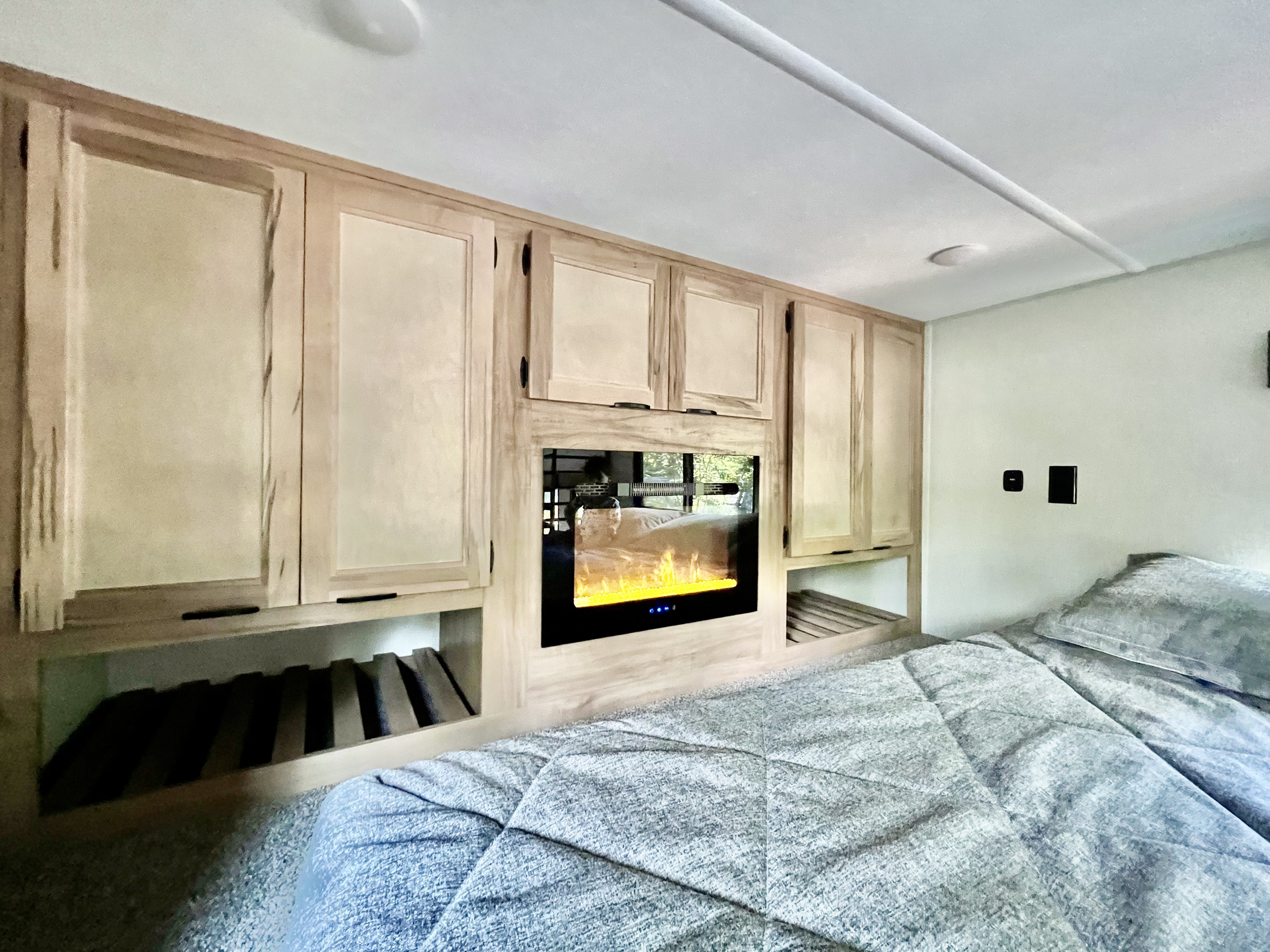 RV rental bedroom with light wood cabinets, an electric fireplace, and slatted storage over a comfortable bed.. Forest River Timberwolf Destination 16ML 2024