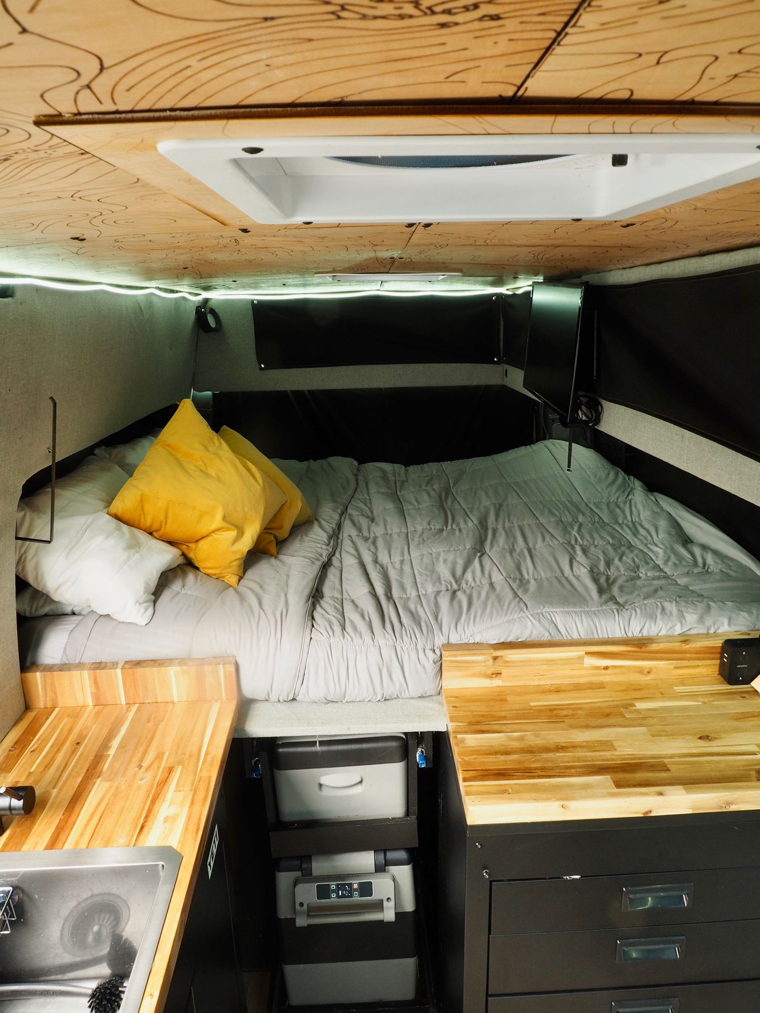 Inside a cozy RV rental: features a comfy bed with yellow pillows, wooden counter, sink, and smart storage.. Mercedes Sprinter 2016