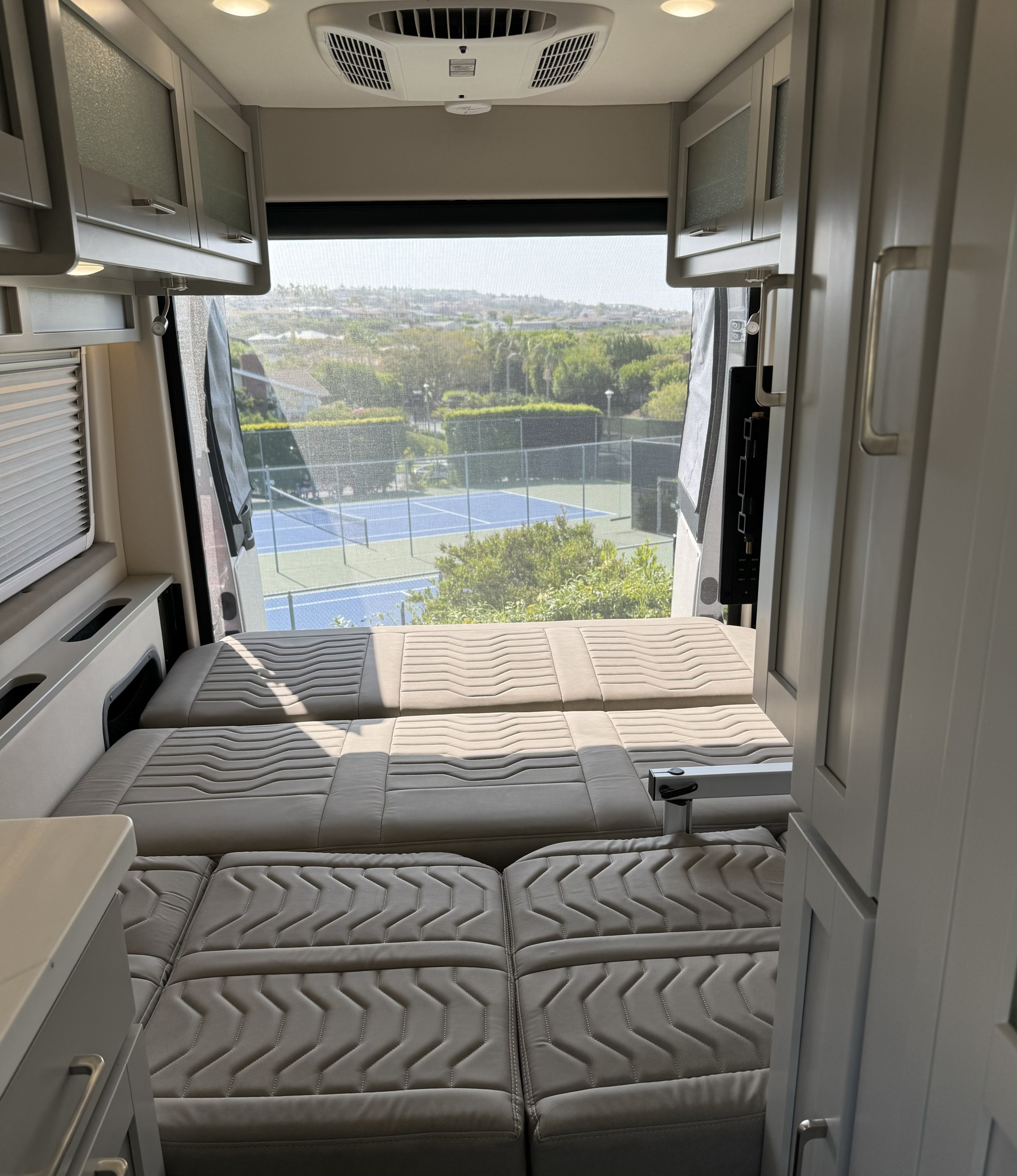 Bright rv rental interior: convertible bed/seating, modern cabinets, and a wide view of tennis courts through the rear window.. RAM Coachmen Nova 2024