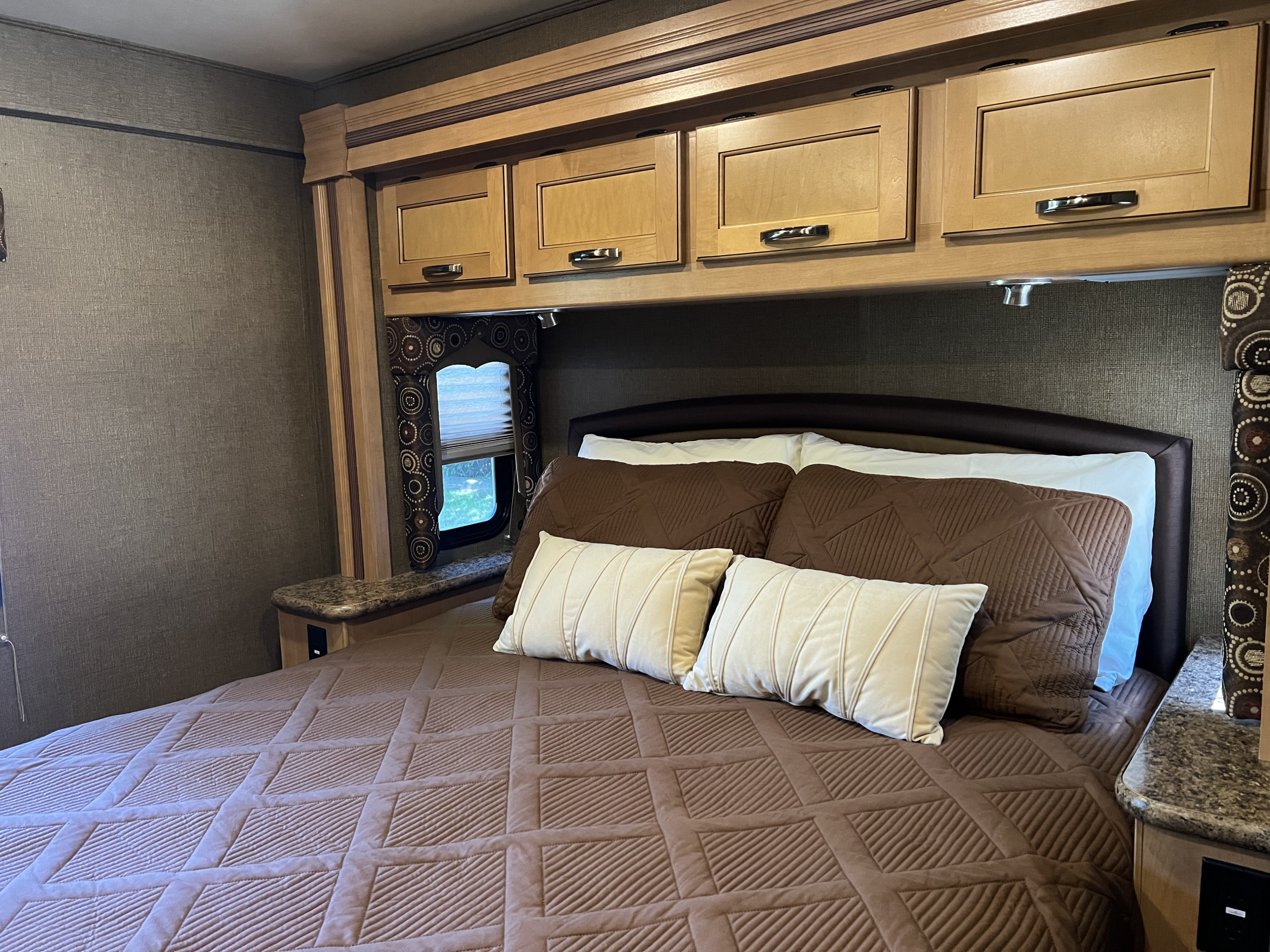Experience comfort in this RV rental bedroom: brown & cream bedding, overhead cabinets, window, and side tables.. Thor Four Winds 2016