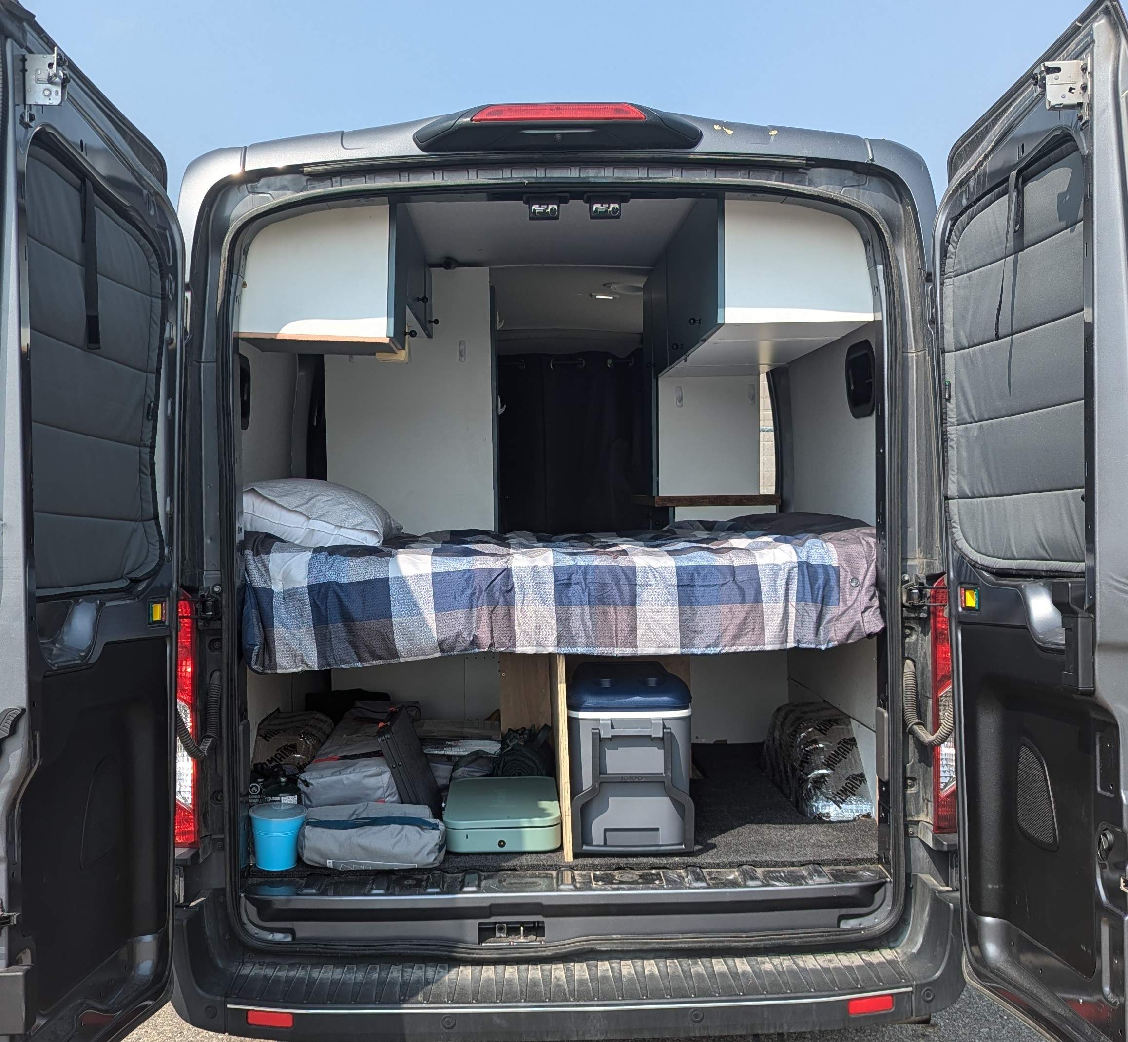 Inside an RV rental van: cozy bed, overhead cabinets, and ample under-bed storage with cooler & stove.. Ford Transit 2022