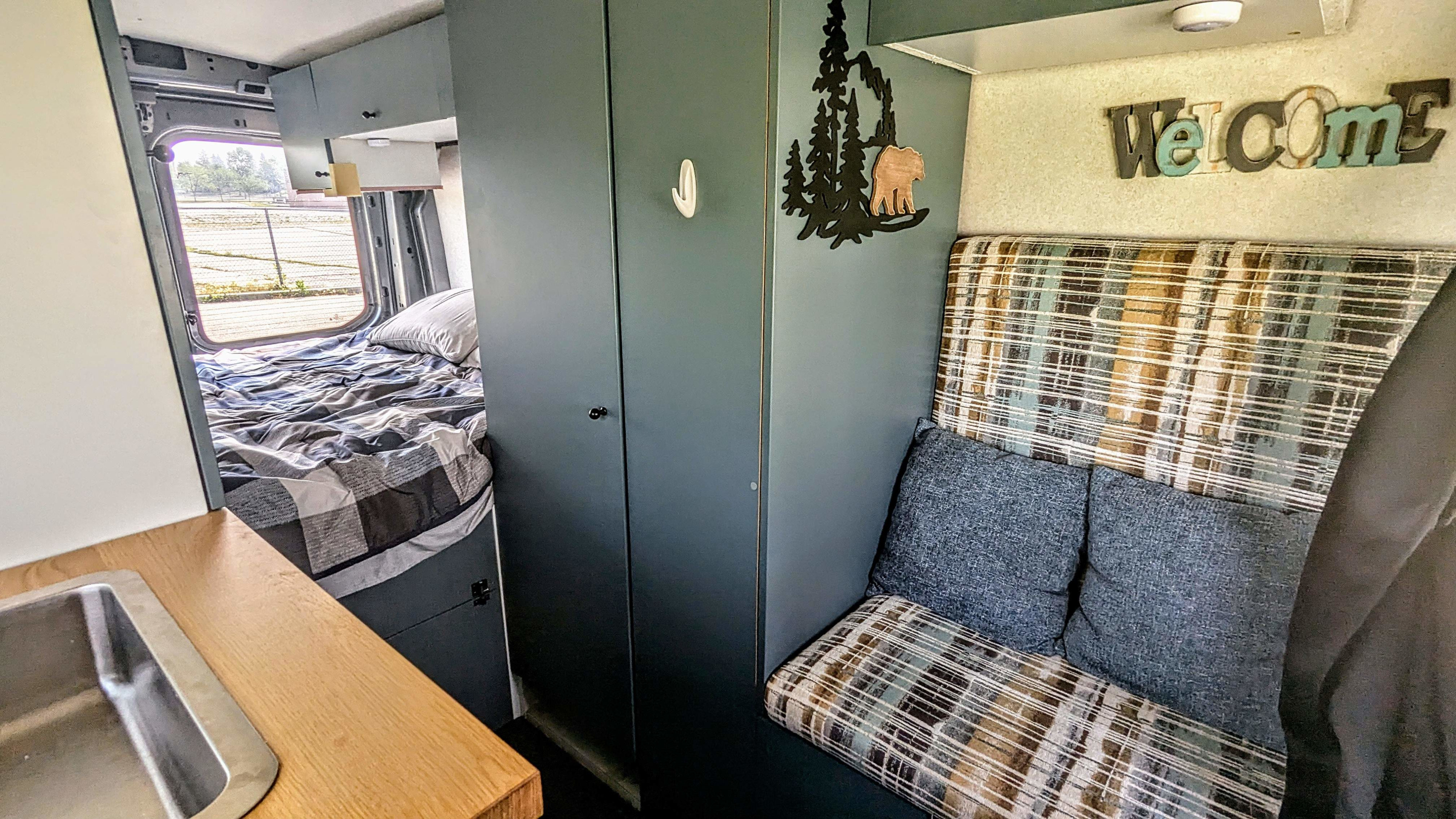 Compact RV rental interior: cozy bed, small kitchen with sink, and a comfortable patterned seating area in a camper van.. Ford Transit 2022