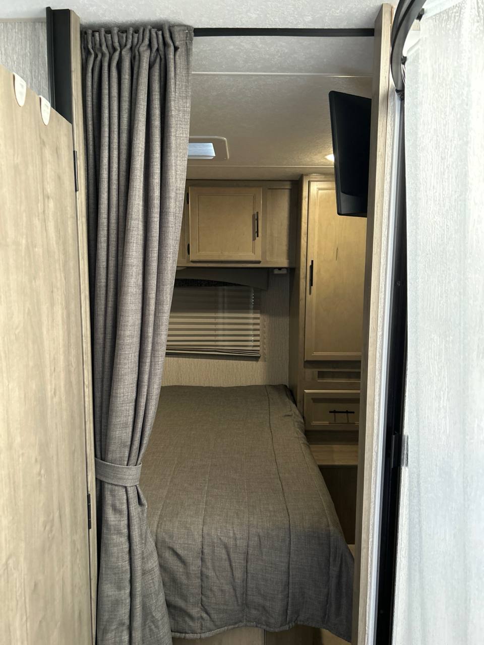 Inside an **RV rental**: a compact bedroom with a gray bed, overhead cabinets, and a wall-mounted TV.. Coachman Freelander 2022