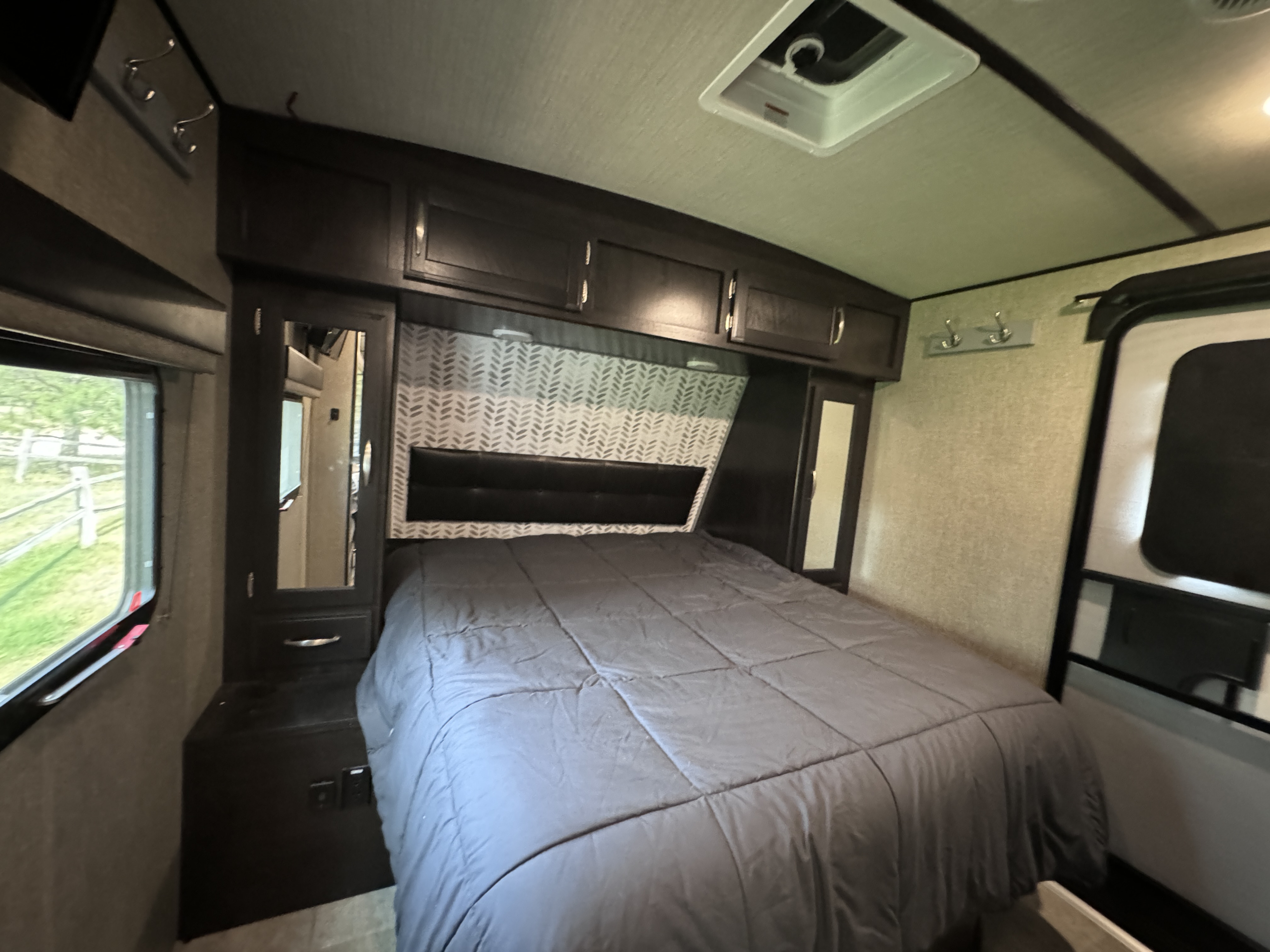 An inviting RV rental bedroom with a gray bed, dark wood cabinets, and a window providing natural light.. Grand Design Imagine 2020