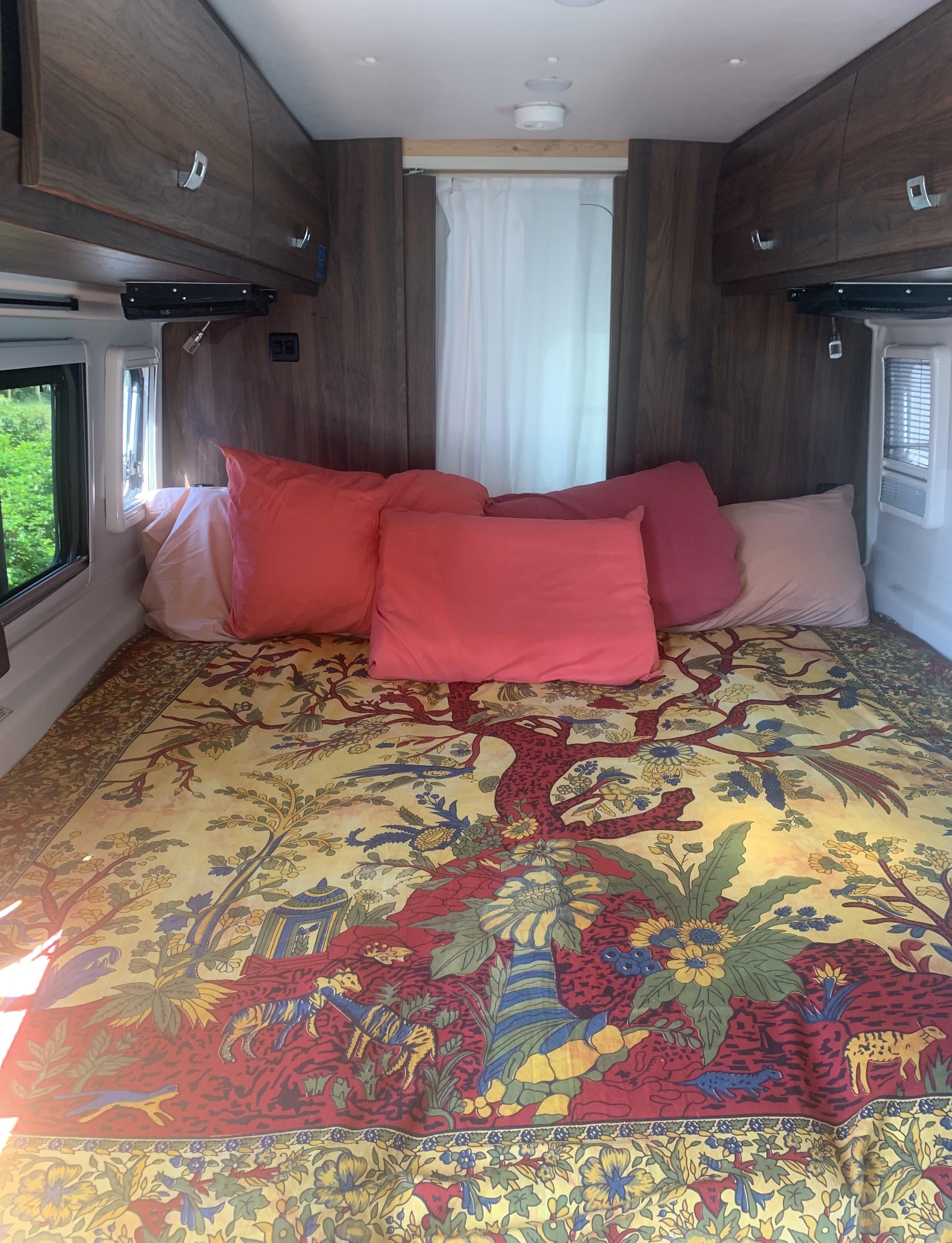 Inside an RV rental: a vibrant bed with an ornate tapestry, plush pillows, and built-in wood cabinets.. Winnebago Travato 2022