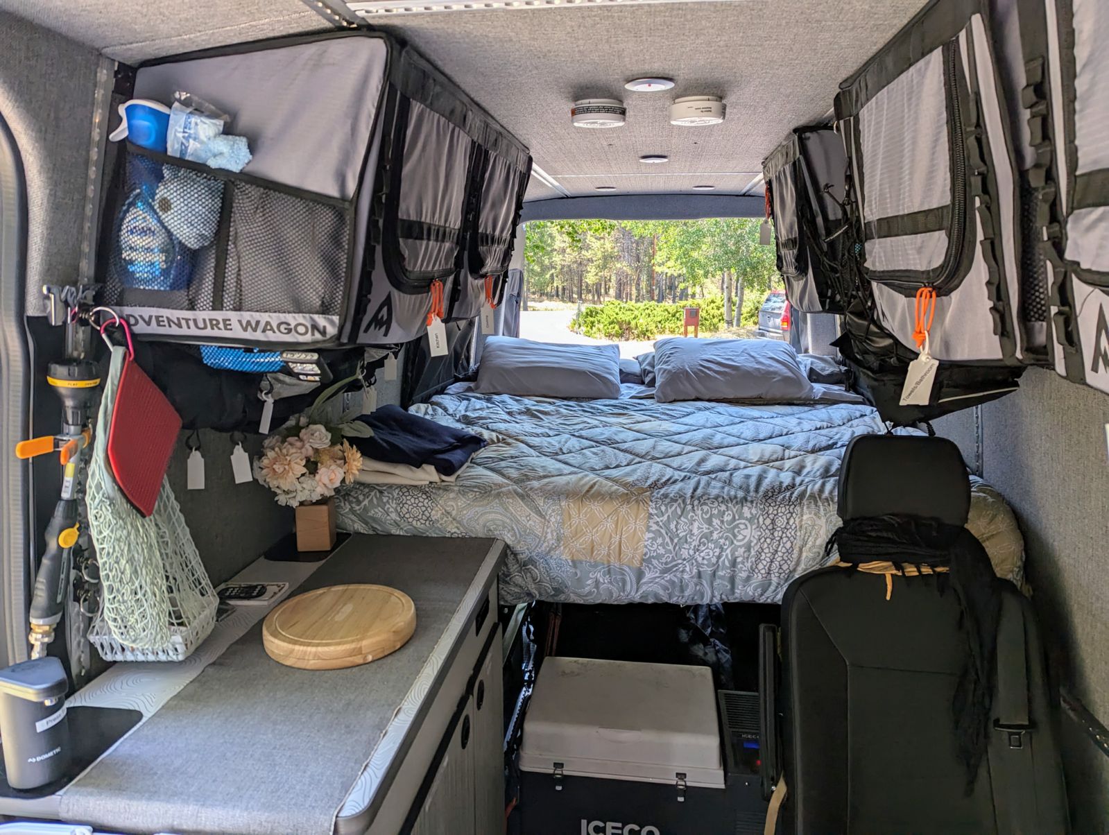 Inside an RV rental: a cozy bed flanked by overhead storage, a compact kitchen counter, and an ICECO fridge below.. Winnebago Adventure Wagon 2023