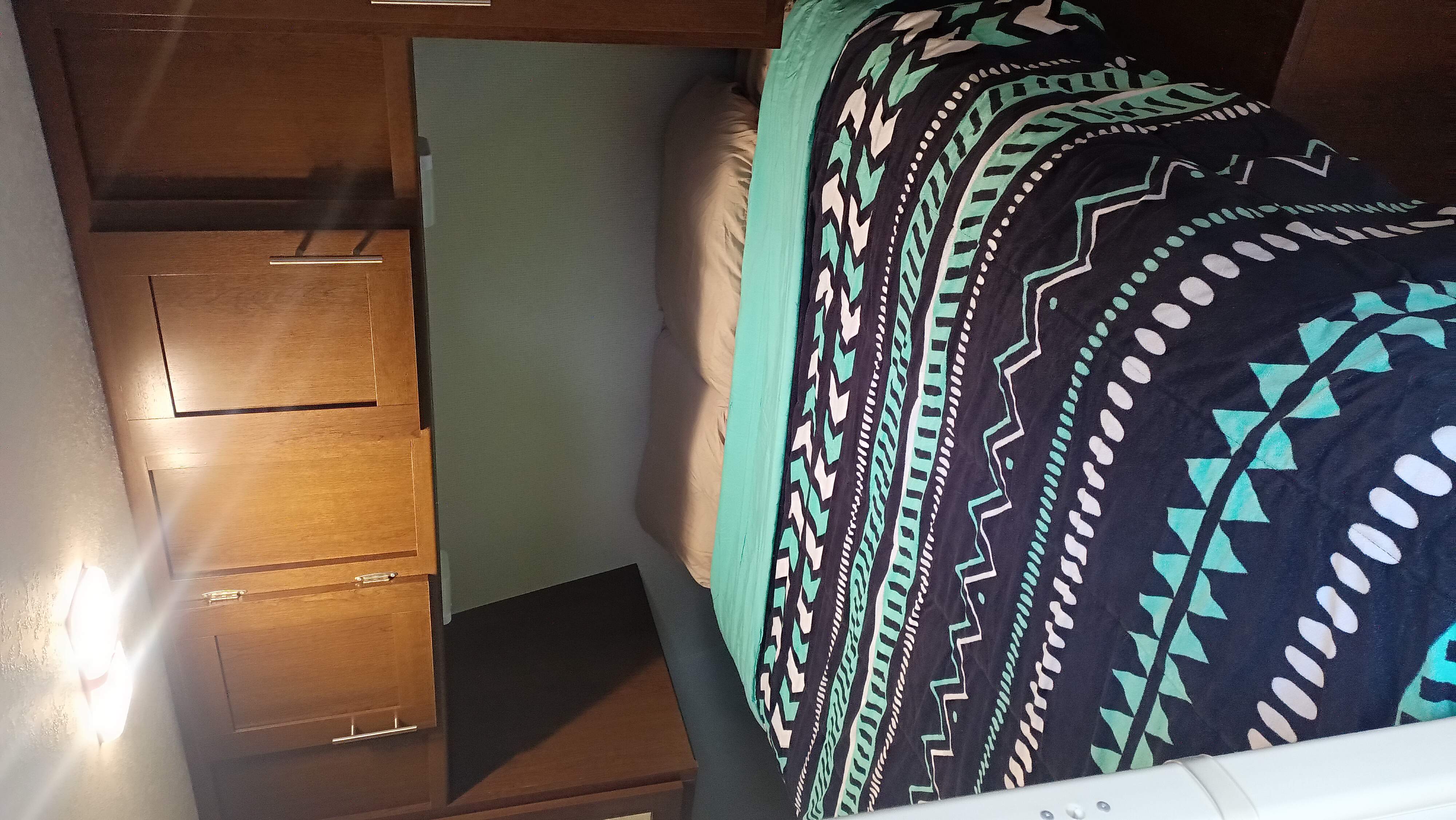 Cozy RV rental bedroom with vibrant black and teal patterned bedding next to wooden cabinets and a bright wall light.. Avenger ATI 2019