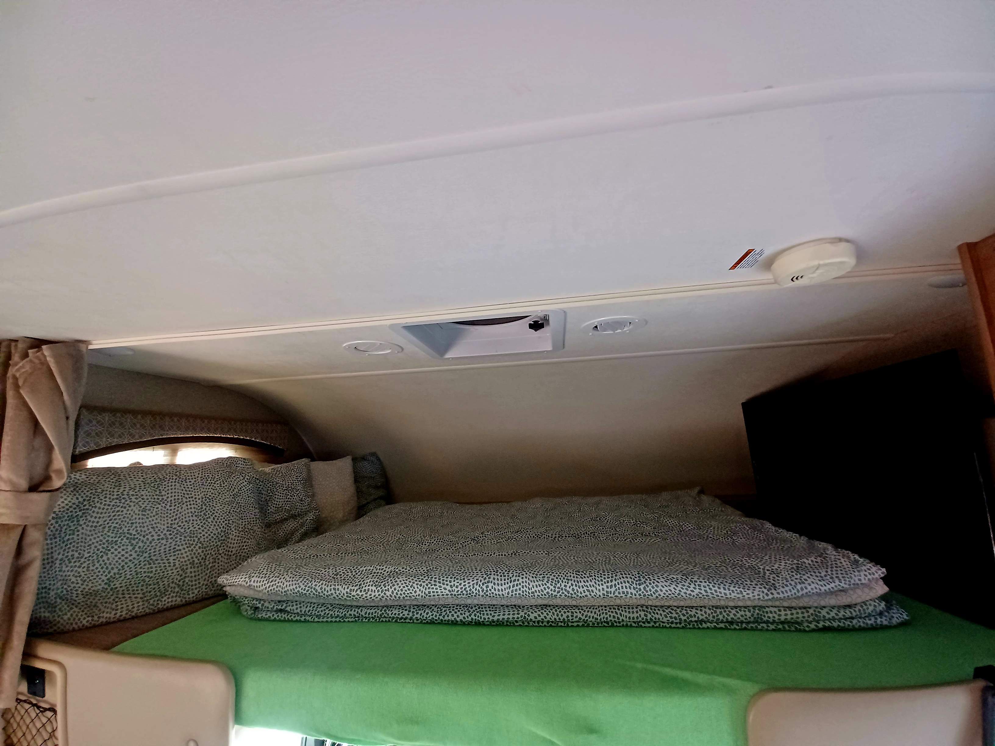 RV rental interior shows a cozy cab-over bed with green sheets, patterned blankets, vents, and a TV. Perfect for road trips.. Thor Chateau 2020