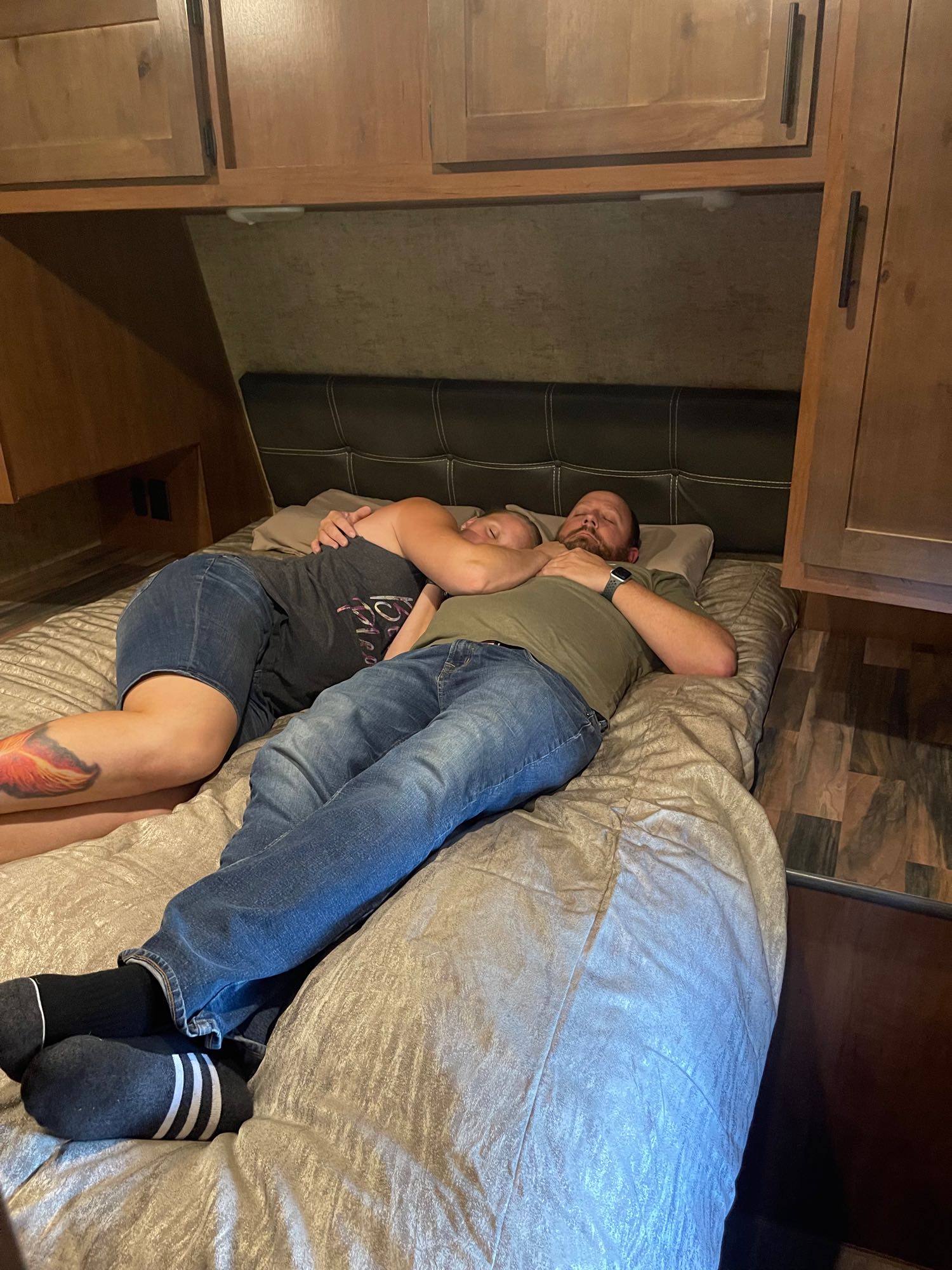A couple, a man and woman, resting comfortably on the bed of their RV rental. Wooden cabinetry surrounds the cozy sleeping area.. Outdoors RV Timber Ridge 2021