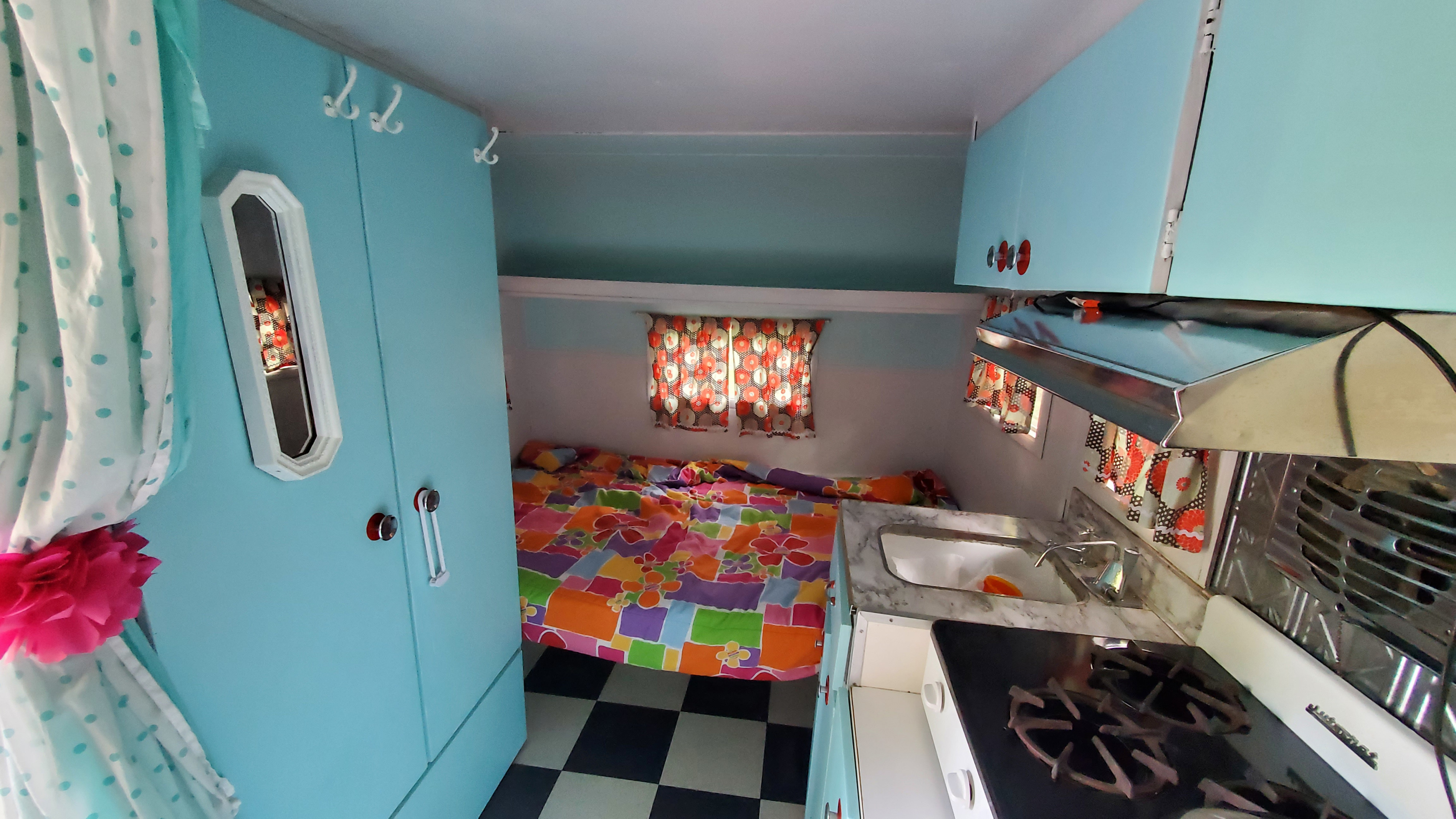 Retro rv rental interior with bright blue walls, colorful bedding, a compact kitchen, and checkered floor.. Skyline Layton 1968
