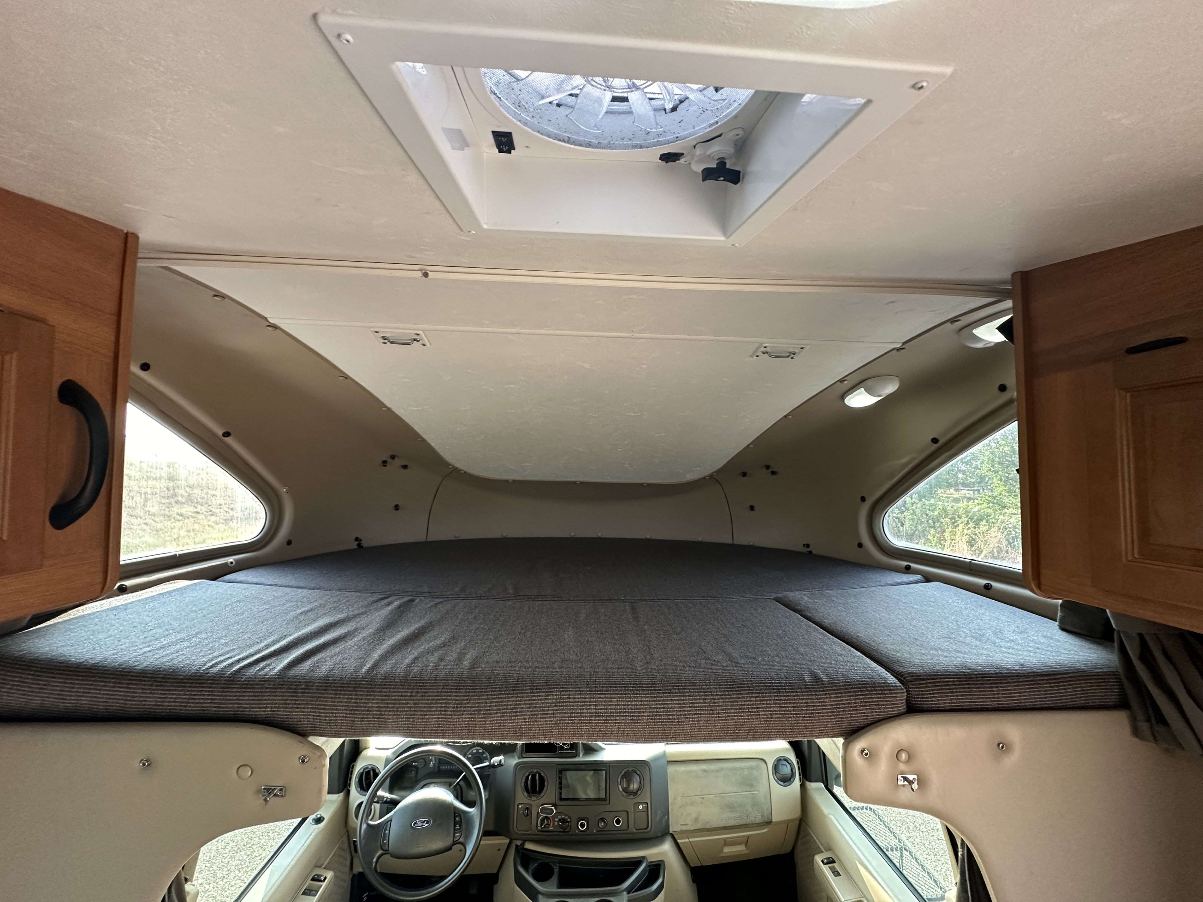 Inside an rv rental: an over-cab bunk with striped mattress, ceiling vent, side windows, and driver's area.. Thor Majestic 2013