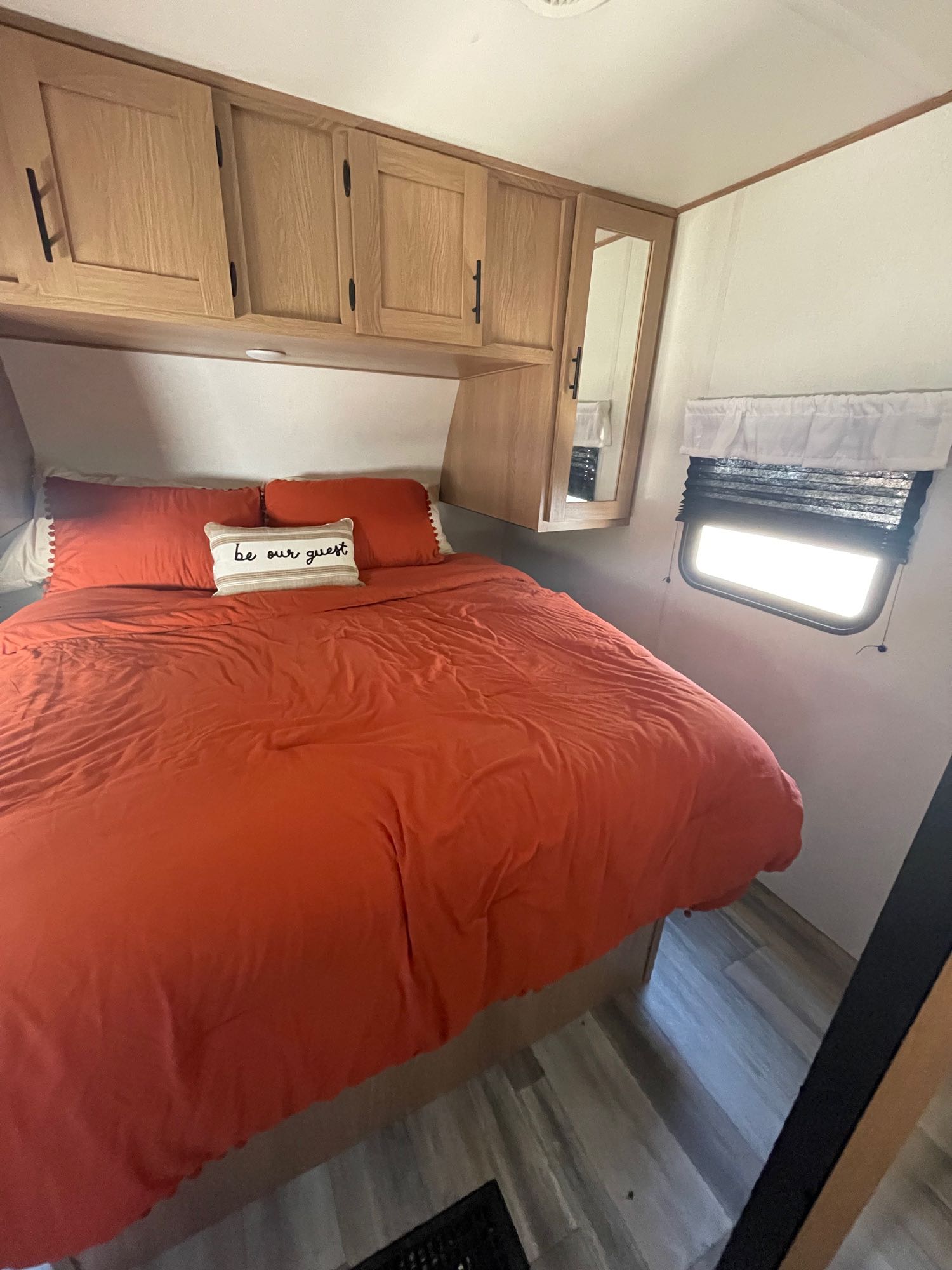 Inside a comfortable RV rental: A warm bedroom with an orange bed, "Be our guest" pillow, wood cabinets & window.. Forest River Avenger 27DBS 2022