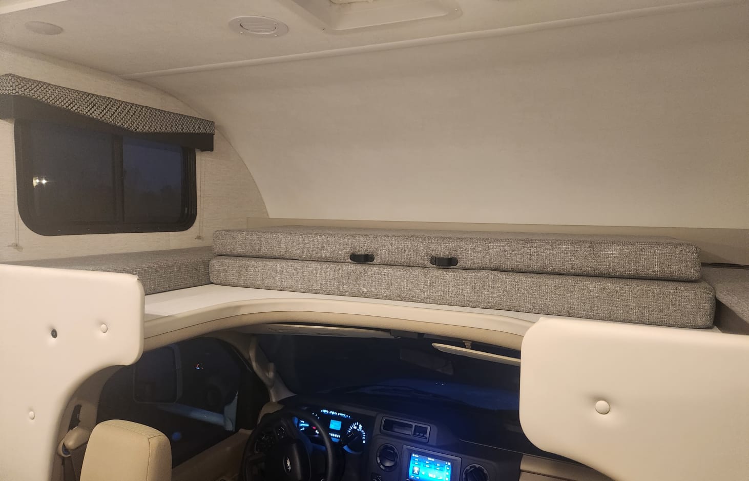This RV rental's interior shows the upper cab-over bunk bed with neatly folded grey cushions and a window, looking down to the driver's area.. Thor Freedom Elite 2024