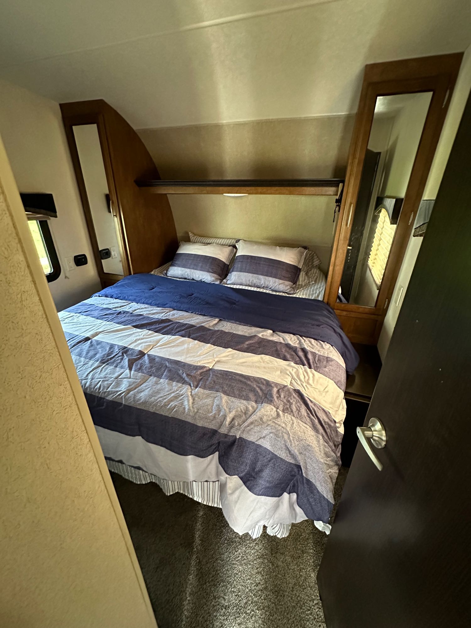 RV rental's cozy bedroom, featuring a blue and white striped bed, built-in mirror, and small window.. Forest River Shockwave 2017