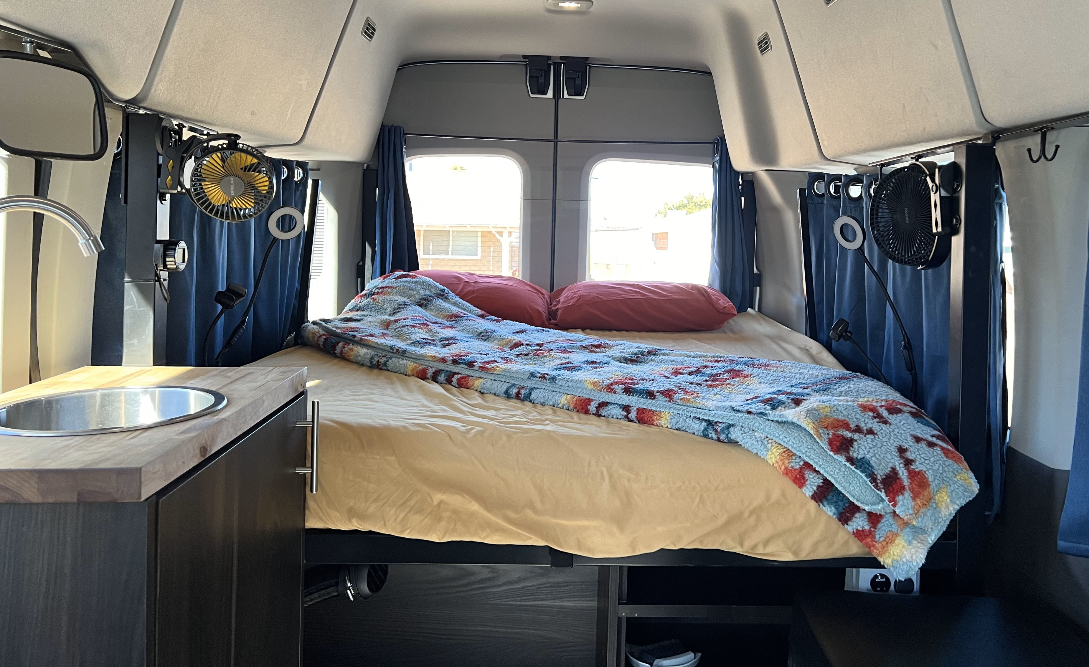 Cozy RV rental interior with bed, built-in sink, fans, and storage for comfortable travel living.. Ford Transit Wagon 2019