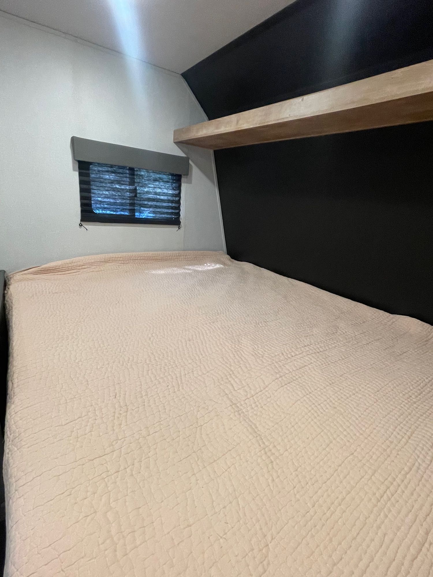RV rental bedroom featuring a cozy, textured bed, window with blinds, and modern black wall with a wooden shelf.. Keystone RV Springdale 2022