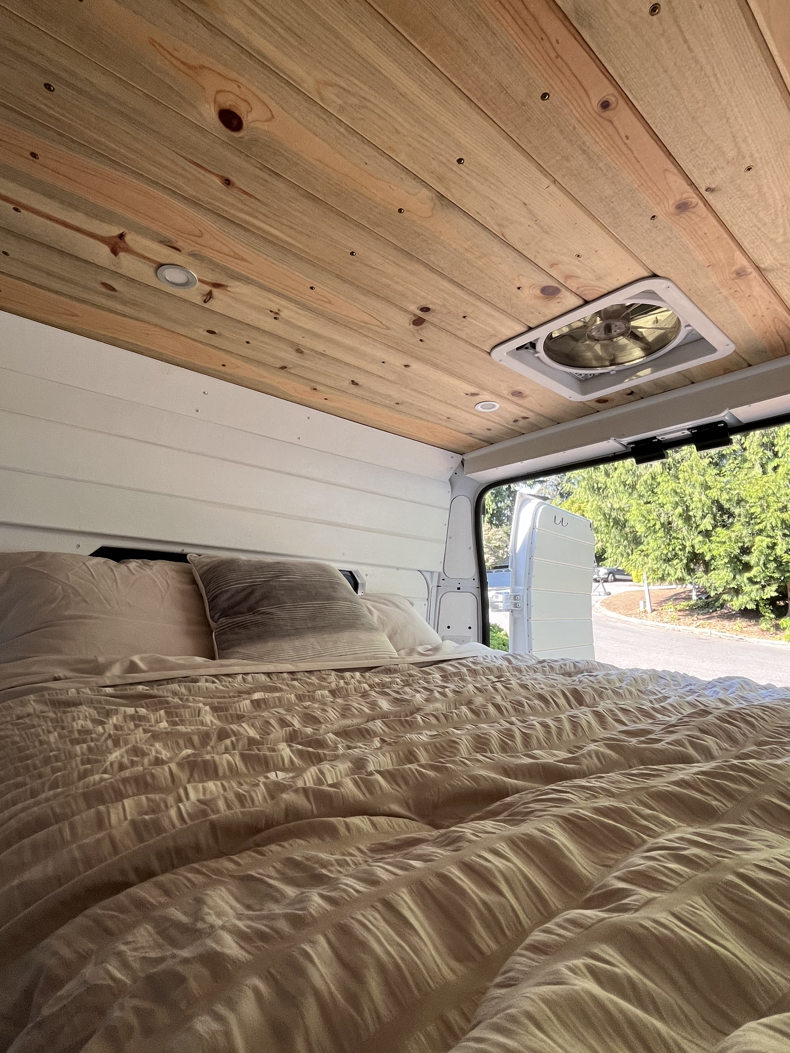 Cozy RV rental bedroom with wood ceiling, fan, soft bed, and open back doors revealing a tree-lined street outside.. Ram Promaster 2500 2021