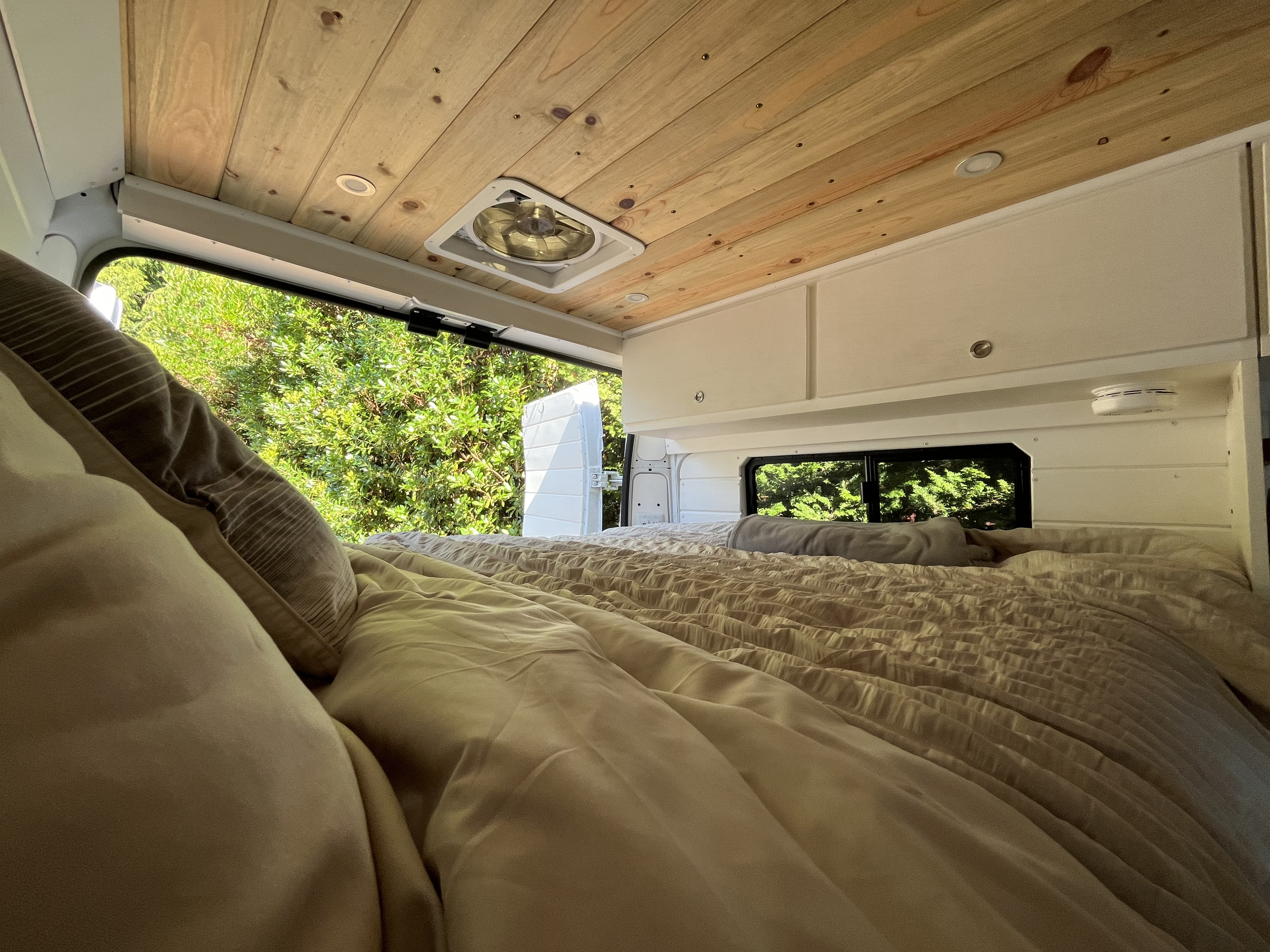 Cozy RV rental camper van interior. Features a comfy bed, wood ceiling, and serene green nature view.. Ram Promaster 2500 2021