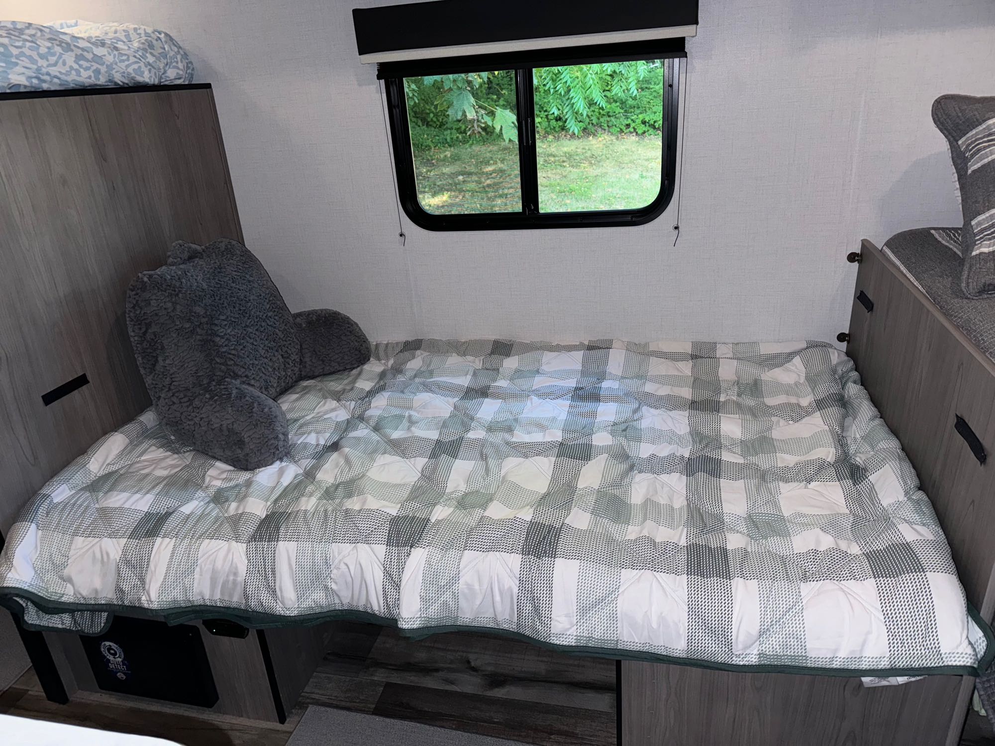 Bright RV rental bedroom: a comfy bed with a plaid quilt, grey backrest pillow, and a window offering a green outdoor view.. Coleman Lantern LT 17B 2024