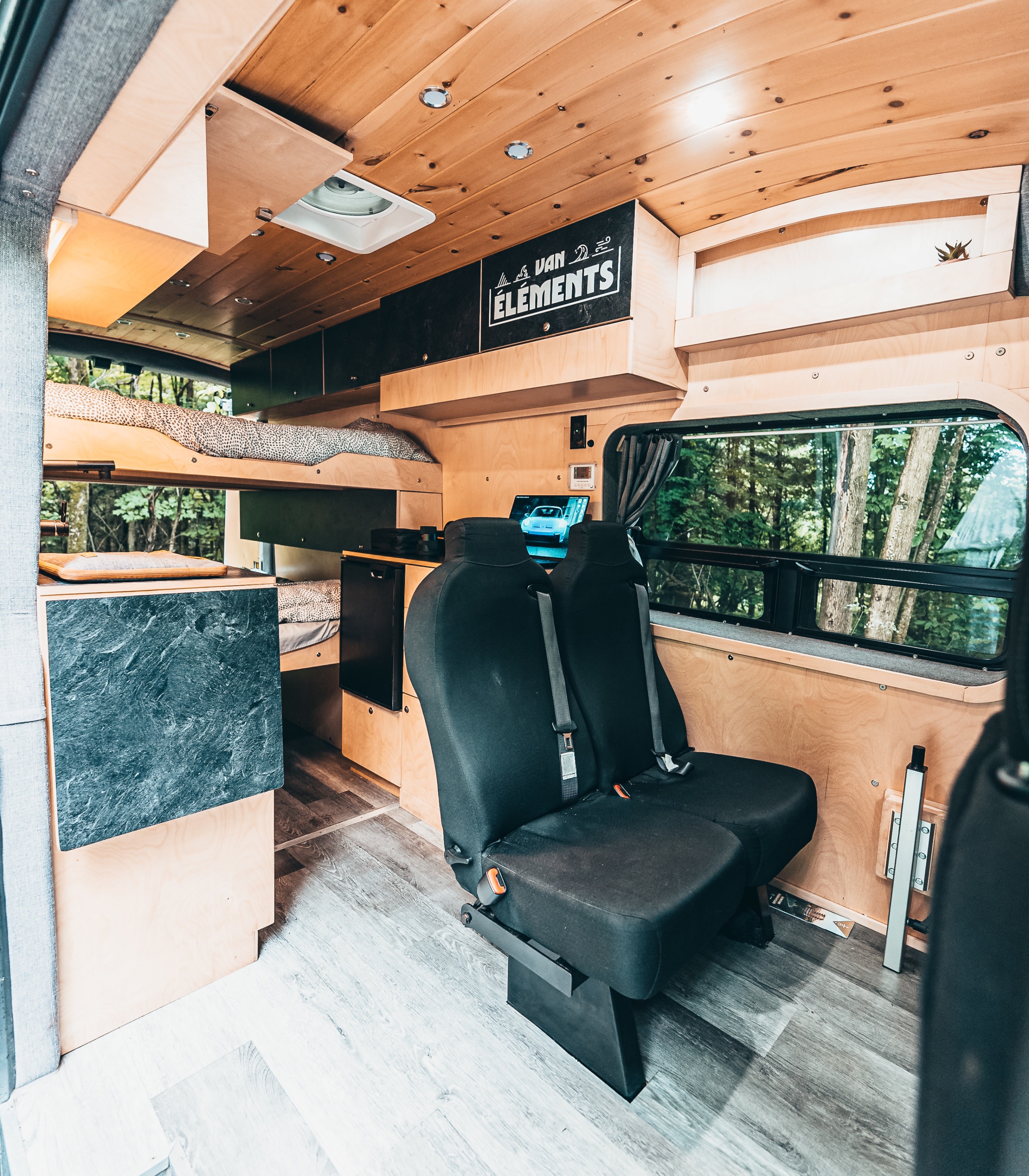 RV rental interior showcasing a custom wooden camper van with bunk beds, passenger seats, and a serene forest view.. Ford Van 2023
