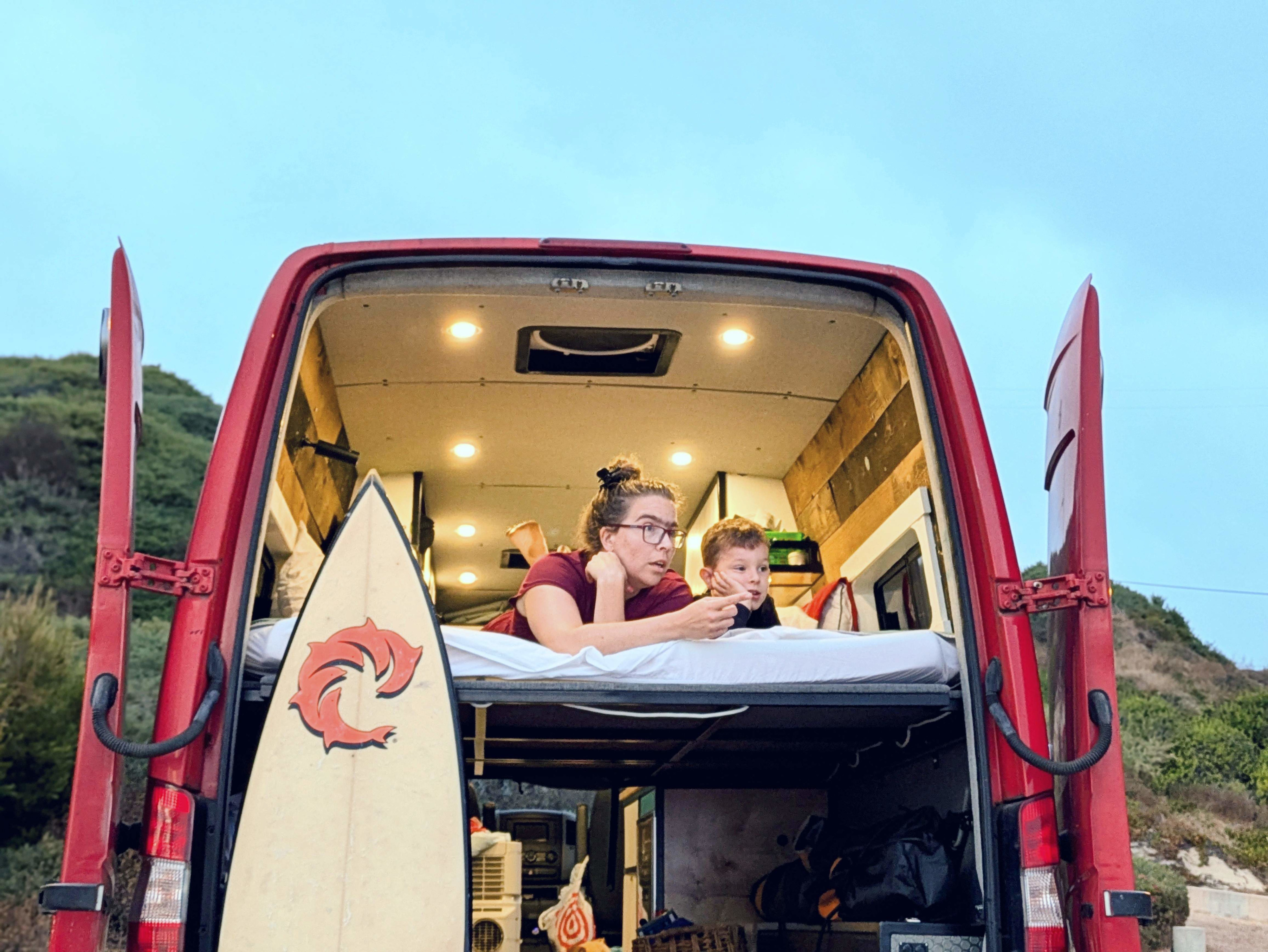 Enjoying van life in an RV rental: A woman and child lounge inside their red camper van, surfboard included. Perfect for family travel.. Mercedes-Benz Sprinter 2008