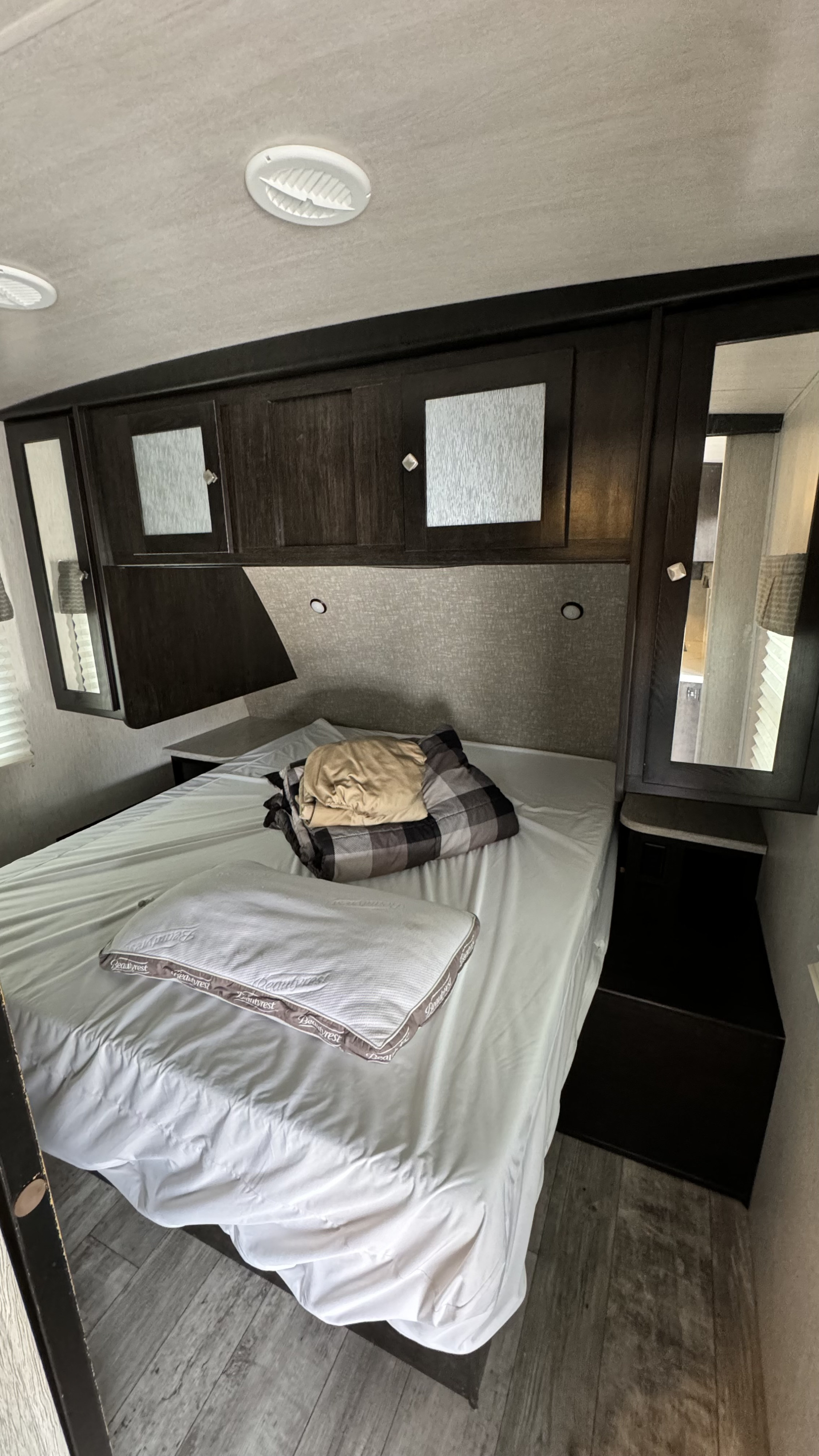 RV rental bedroom with white sheets, dark wood cabinets, and functional ceiling vents, offering a compact sleeping area.. Heartland North Trail 2019