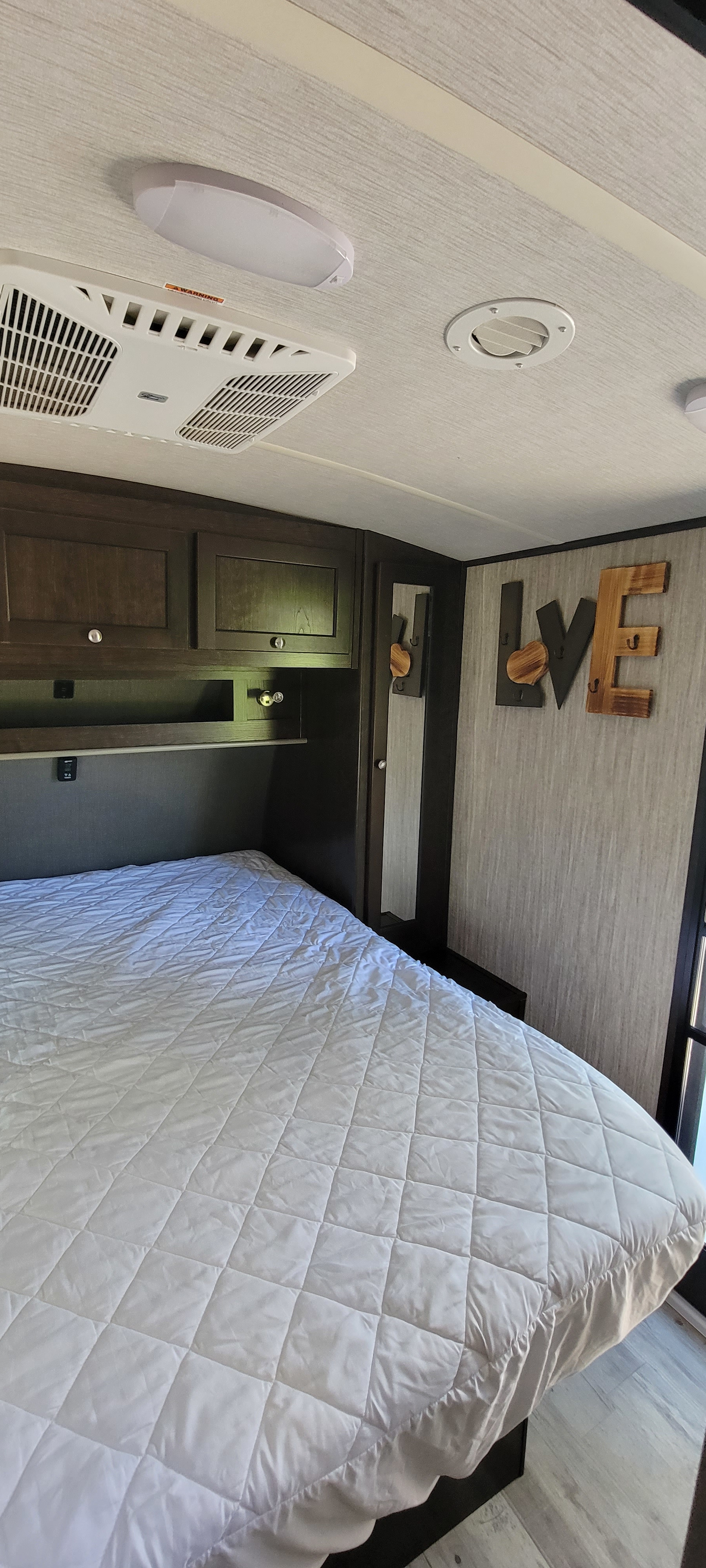 Cozy RV rental bedroom with a quilted white bed, dark wood overhead cabinets, and an AC unit, featuring "LOVE" decor.. Heartland Sundance 2019