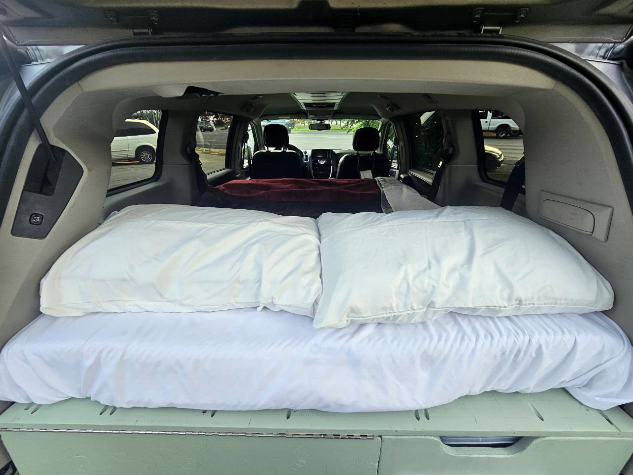 Inside an RV rental: a transformed minivan features a comfy bed with white pillows and a custom storage platform.. Chrysler Town Country 2014