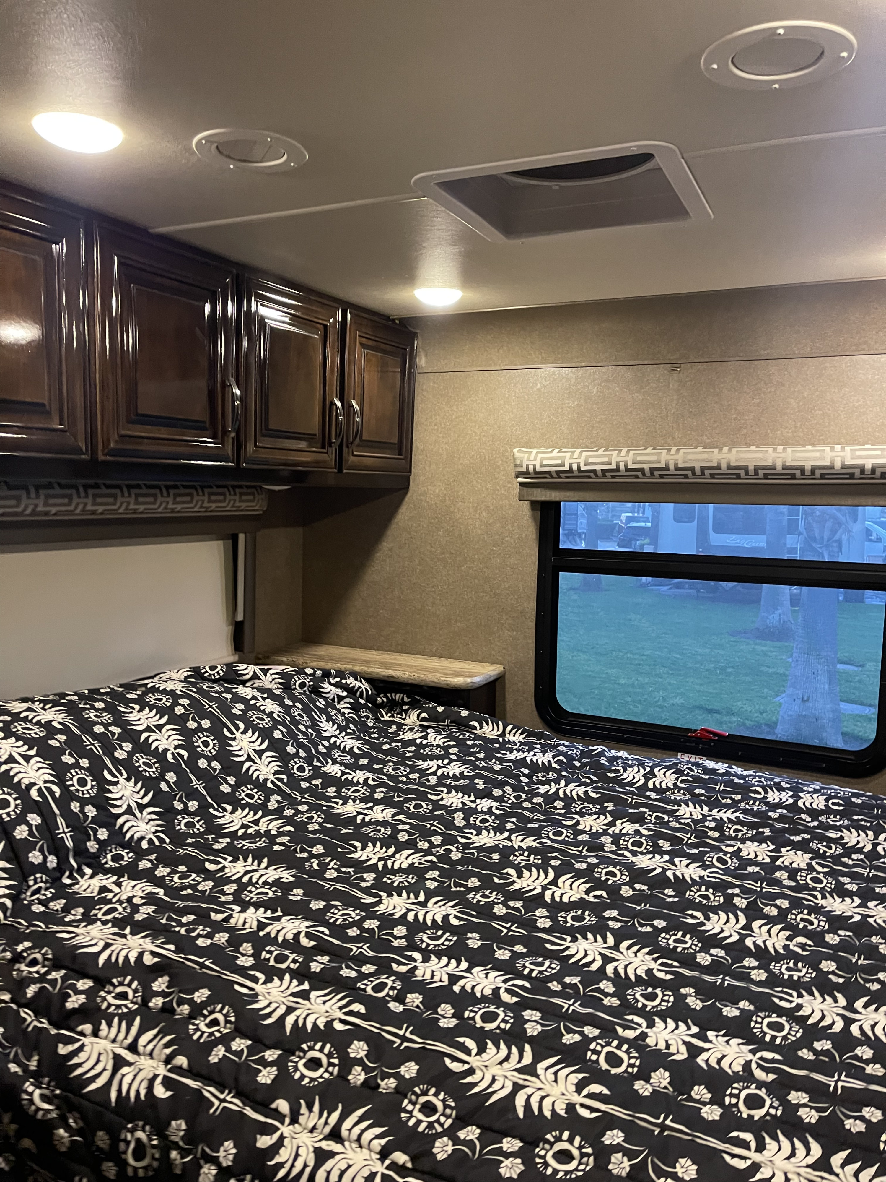 Black and white patterned bed in an RV rental bedroom, with wood cabinets, ceiling lights, and an outdoor window view.. Thor Quantum 2018
