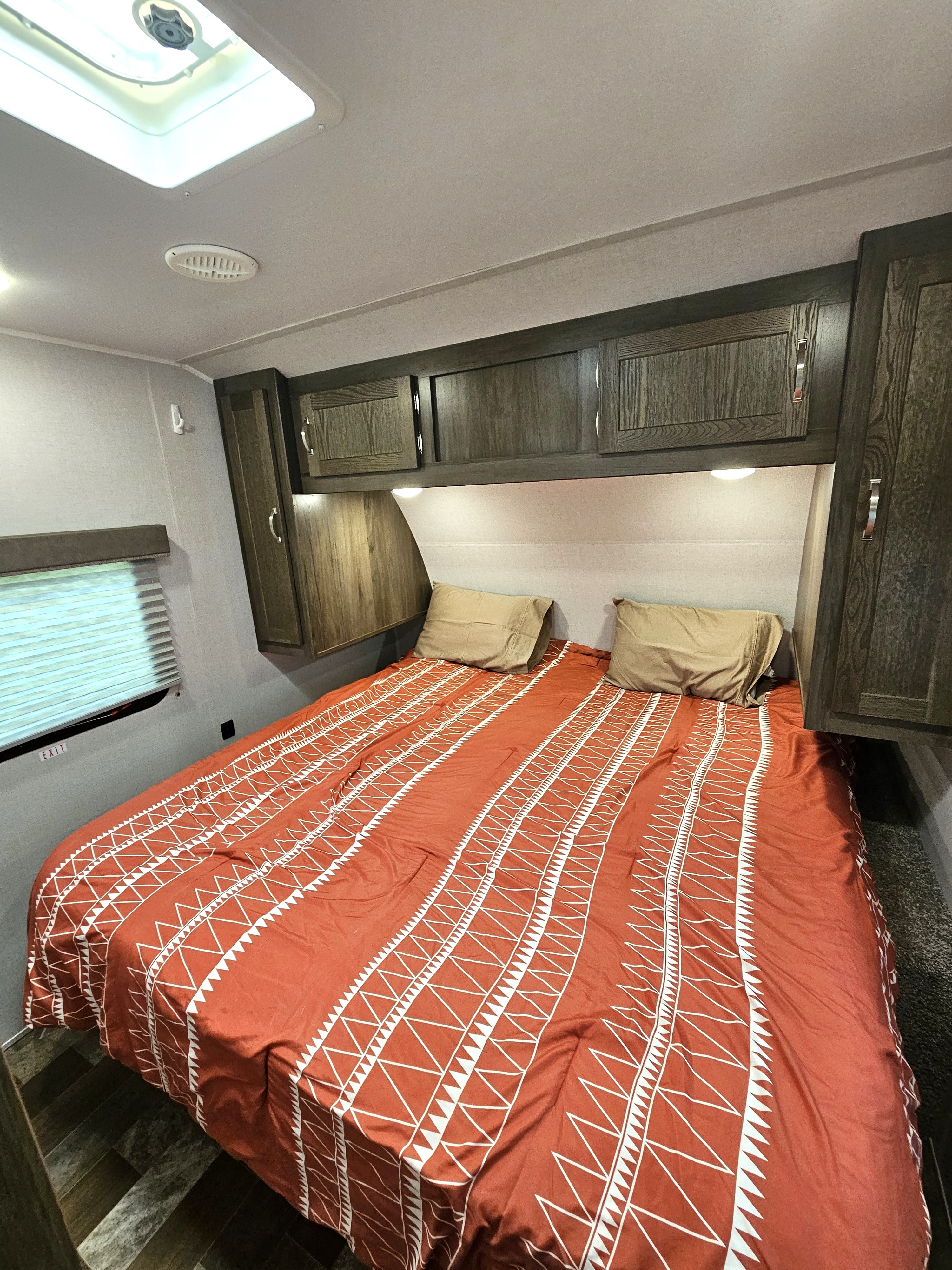 Explore this comfortable RV rental bedroom featuring a spacious bed, overhead cabinets, and window with blinds.. East to West Silver Lake 2021