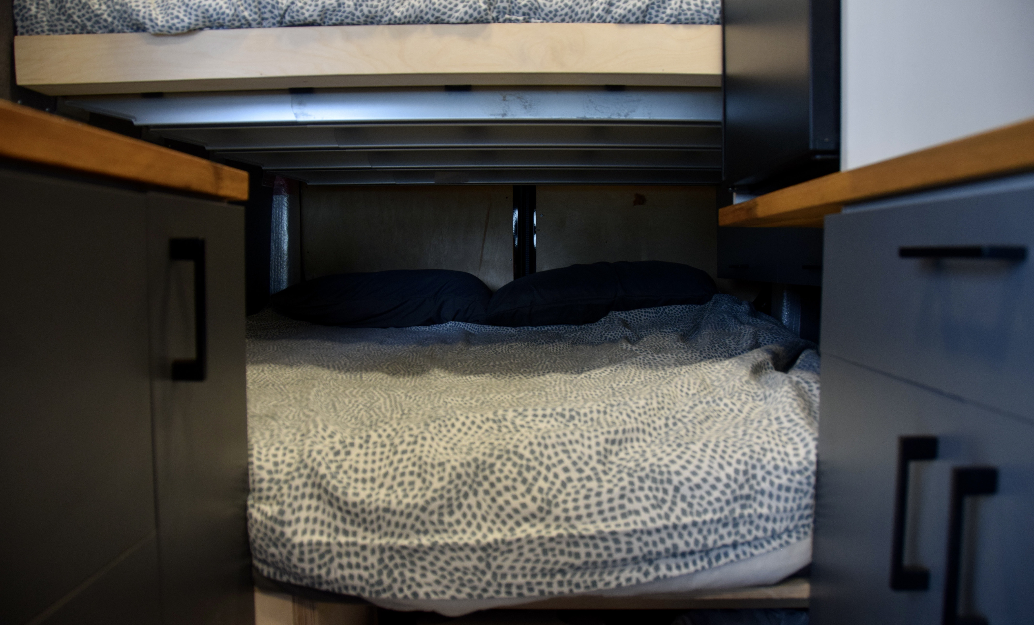 RV rental bunk beds and storage, showing cozy lower sleeping area with pillows and duvet and dark cabinets.. Ford Van 2023