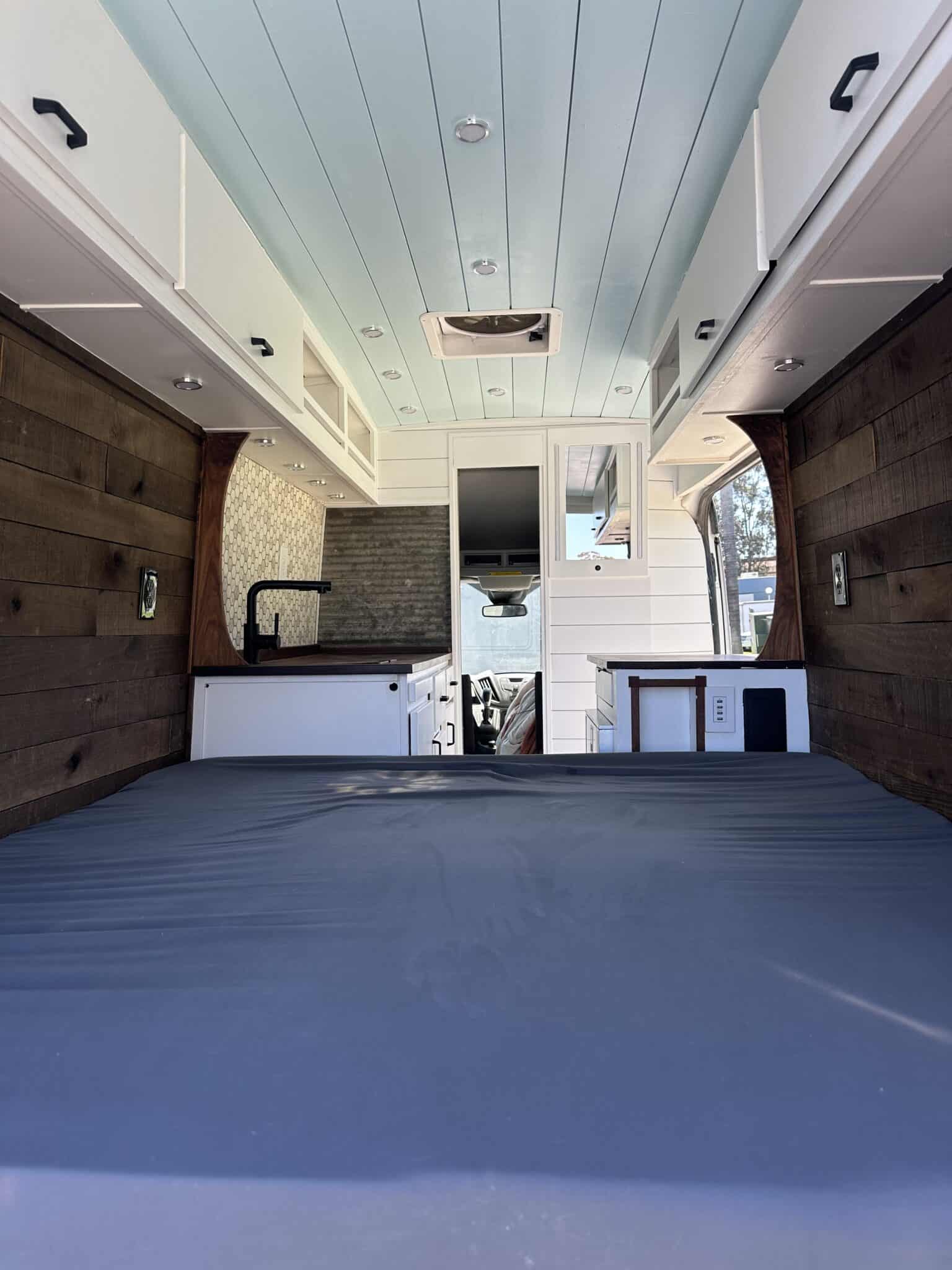 Inside a cozy rv rental camper van: a comfortable bed, full kitchen area, wood-paneled walls, and overhead storage.. Ford Transit 2017