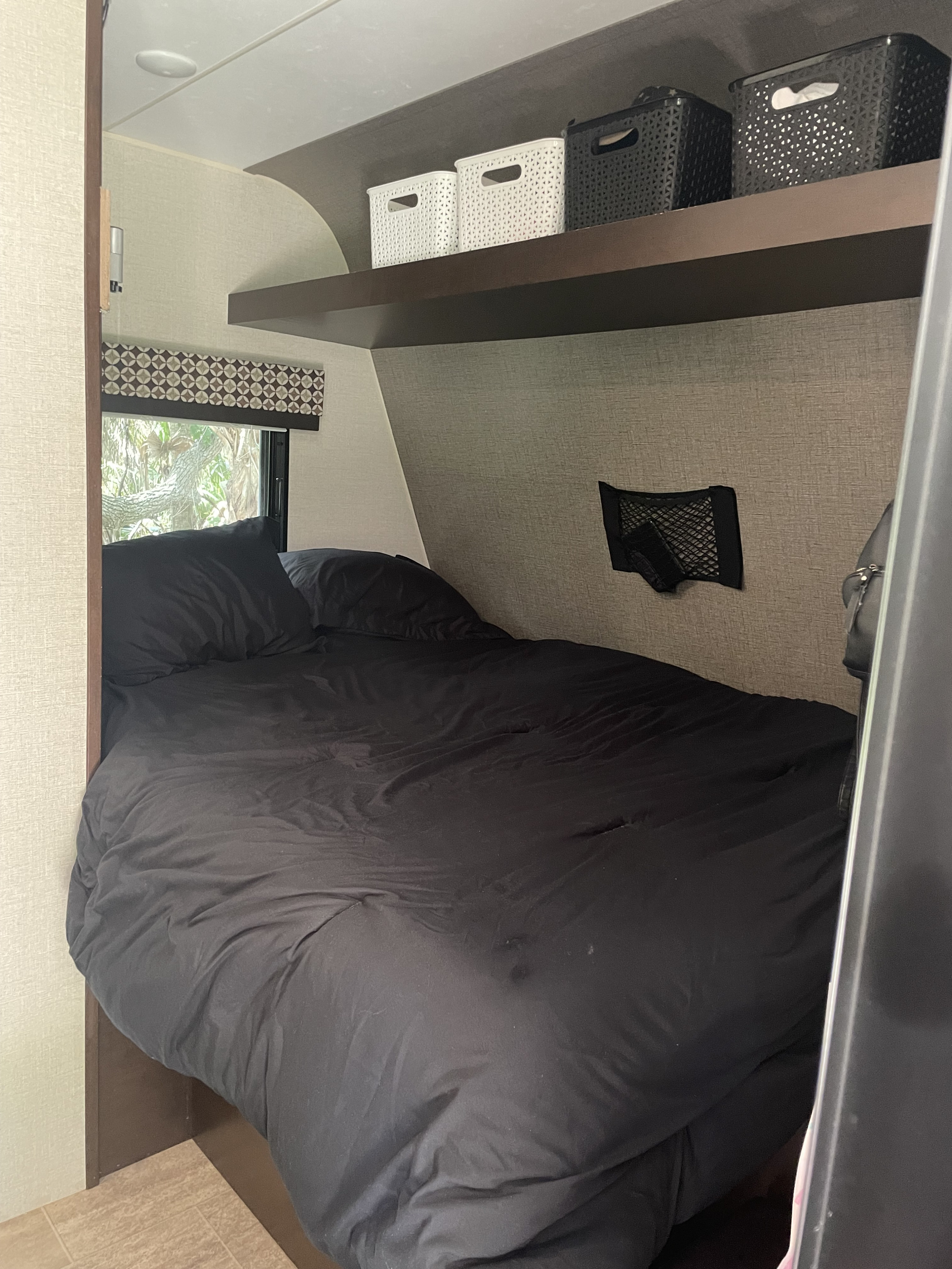 Inside a comfy RV rental: compact bedroom featuring black bedding, a window, an overhead shelf with bins, and a mesh wall pocket.. K-Z Manufacturing Sportsmen 2019