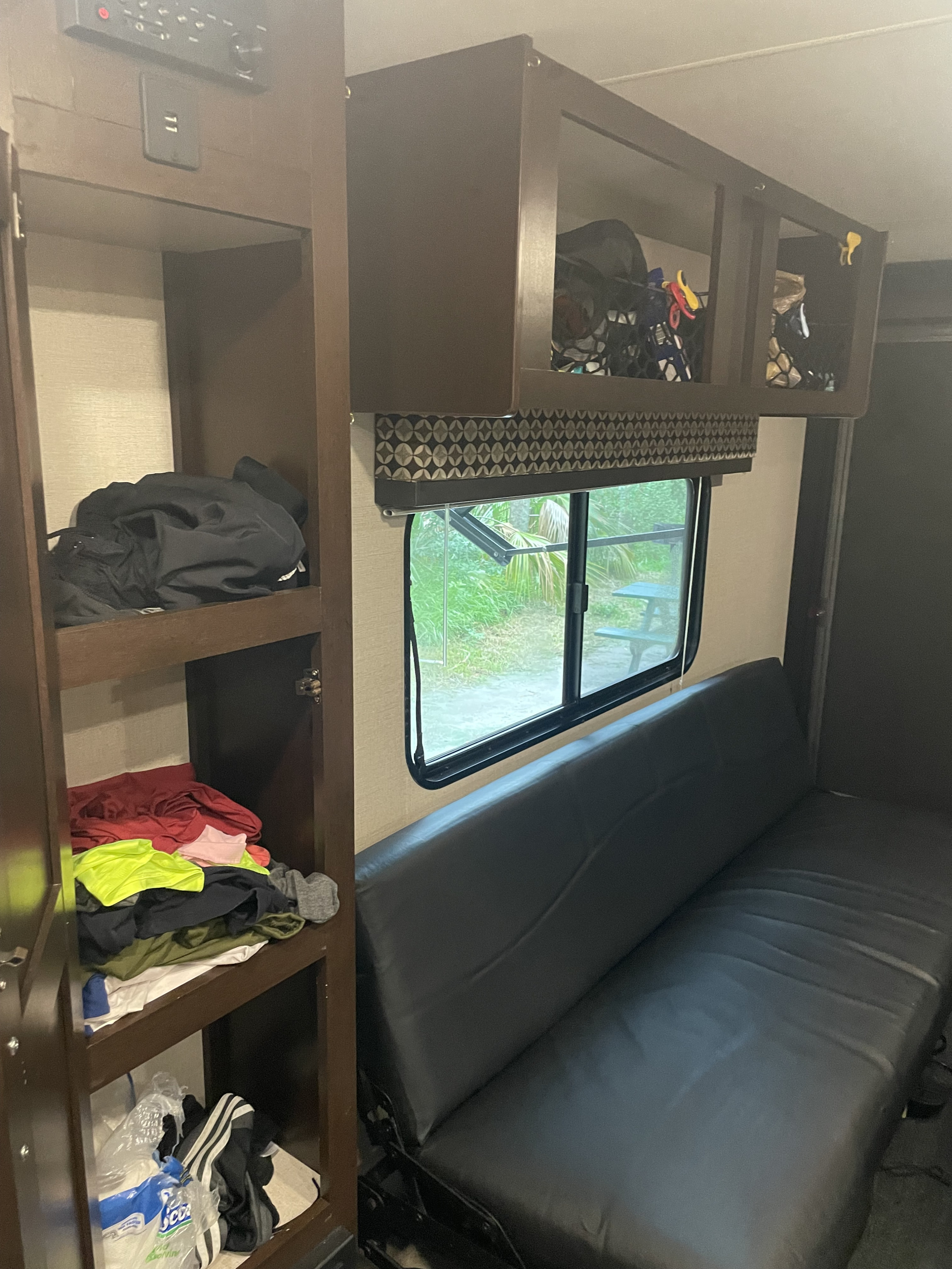 This RV rental interior features practical storage shelves, an inviting convertible couch, and a window overlooking a green campsite.. K-Z Manufacturing Sportsmen 2019