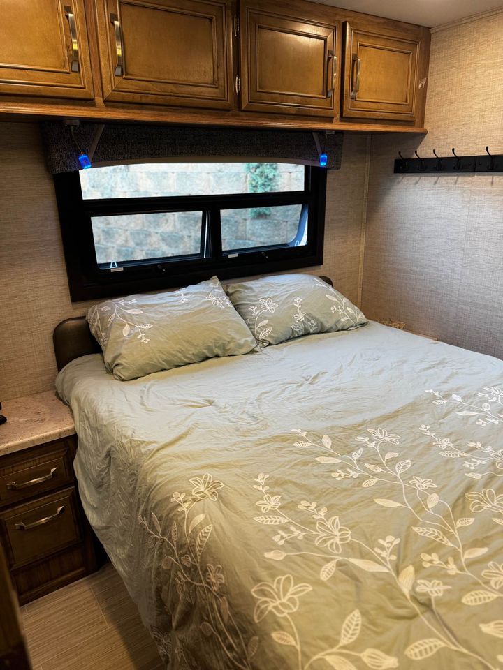 Cozy RV rental bedroom with floral bedding, overhead cabinets, and window view. Perfect for your next adventure.. Jayco Greyhawk 2021